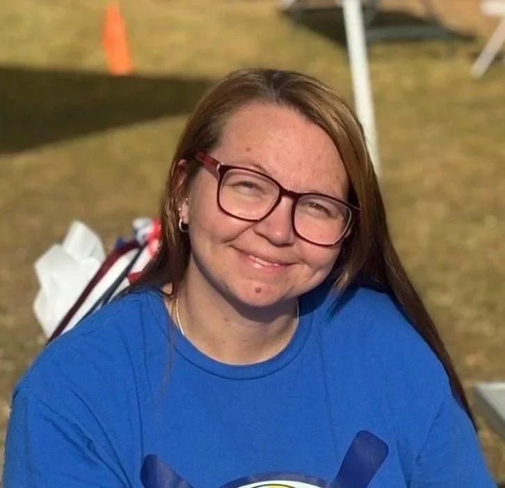 A young woman with long brown hair, glasses, and freckles, smiling outdoors with a tent in the background.