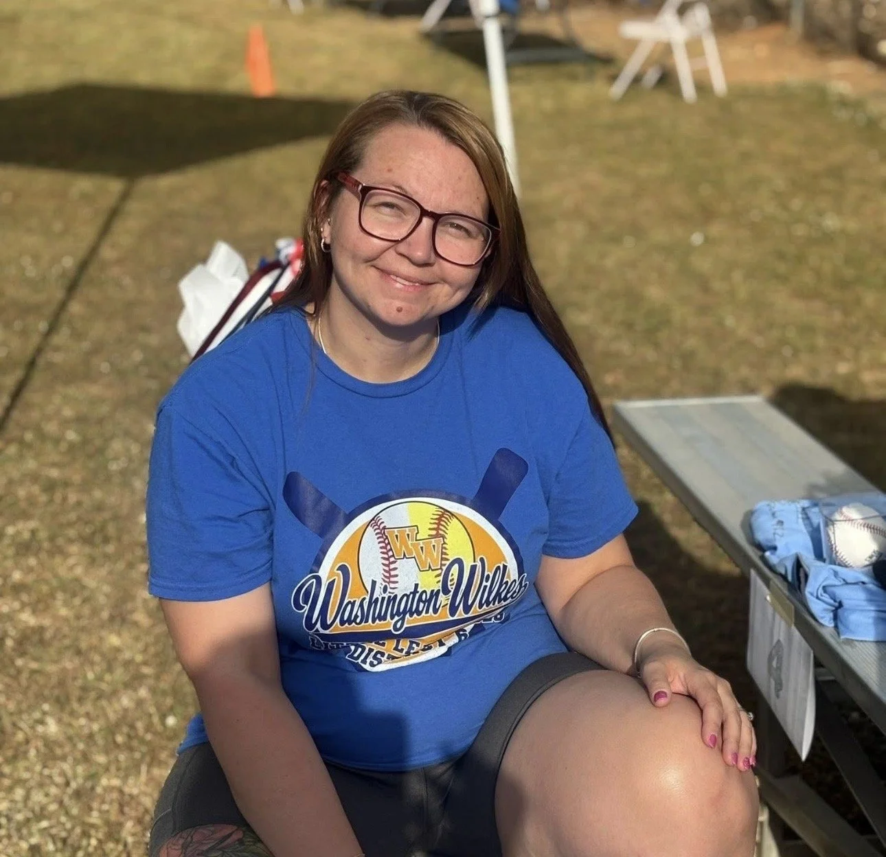 A smiling woman with glasses, wearing a blue Washington Wilkes shirt, sitting outdoors on a sunny day, with a baseball cap and other items on a nearby table.