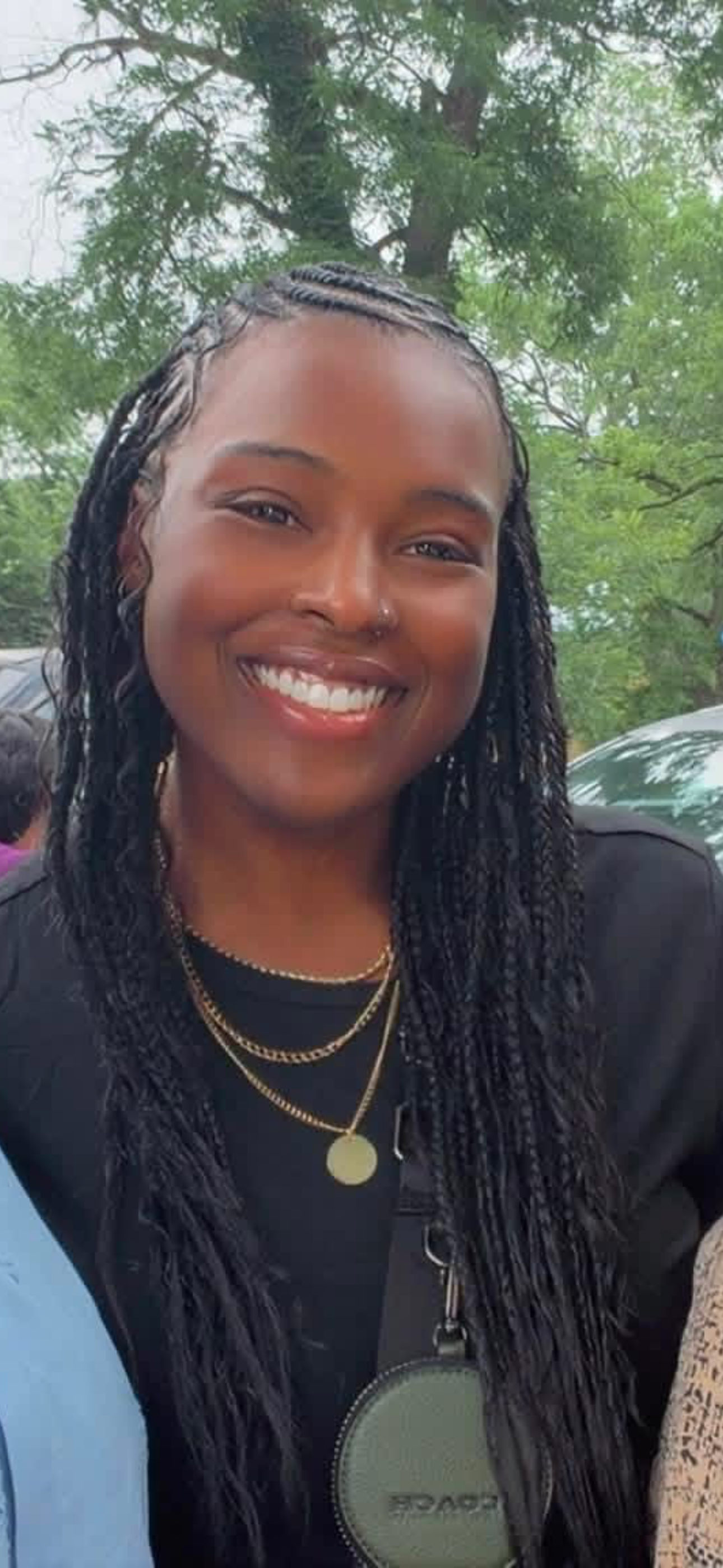 A smiling woman with dark braided hair, wearing layered gold necklaces, a black top, and a black jacket, standing outdoors with green trees in the background.