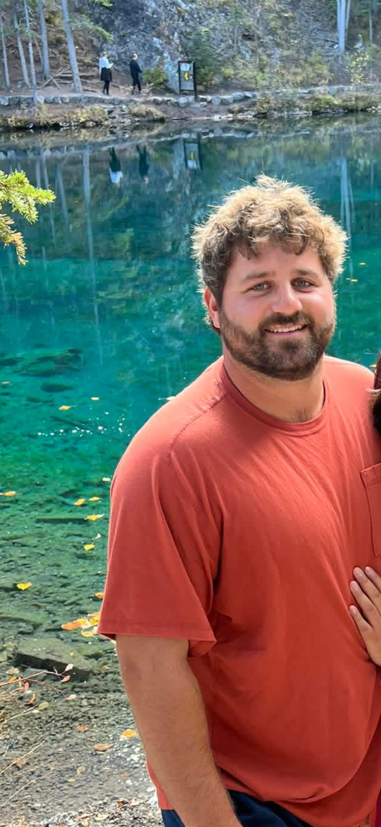 Man in a red t-shirt standing outdoors near a clear blue lake with trees and two people walking on a trail in the background.