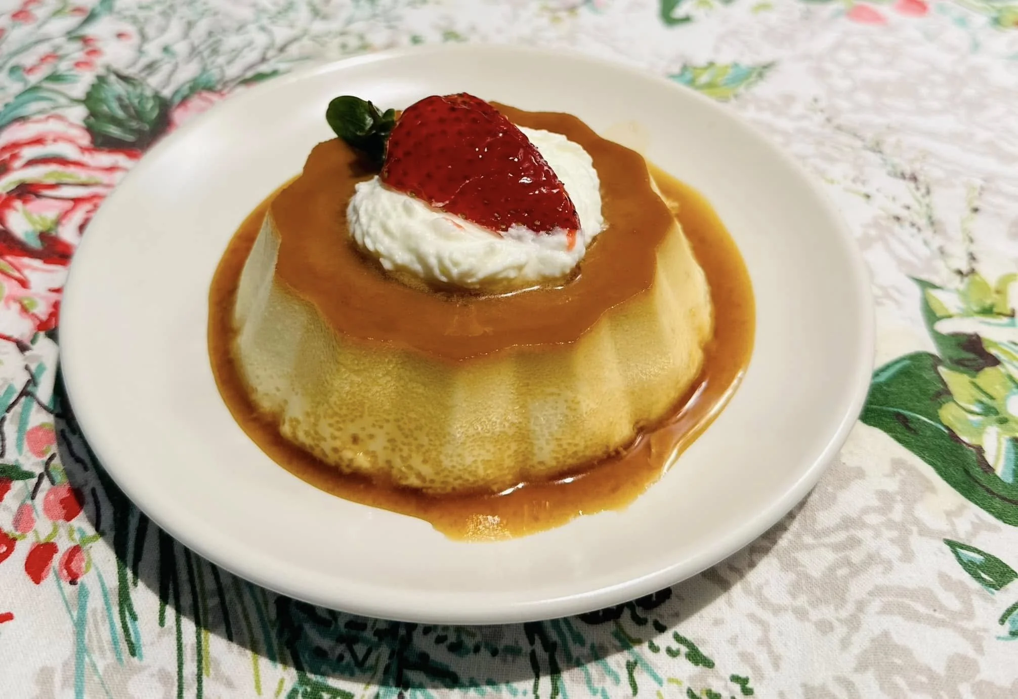 A dessert featuring a flan topped with caramel sauce, whipped cream, a fresh strawberry, and a small green leaf on a white plate.