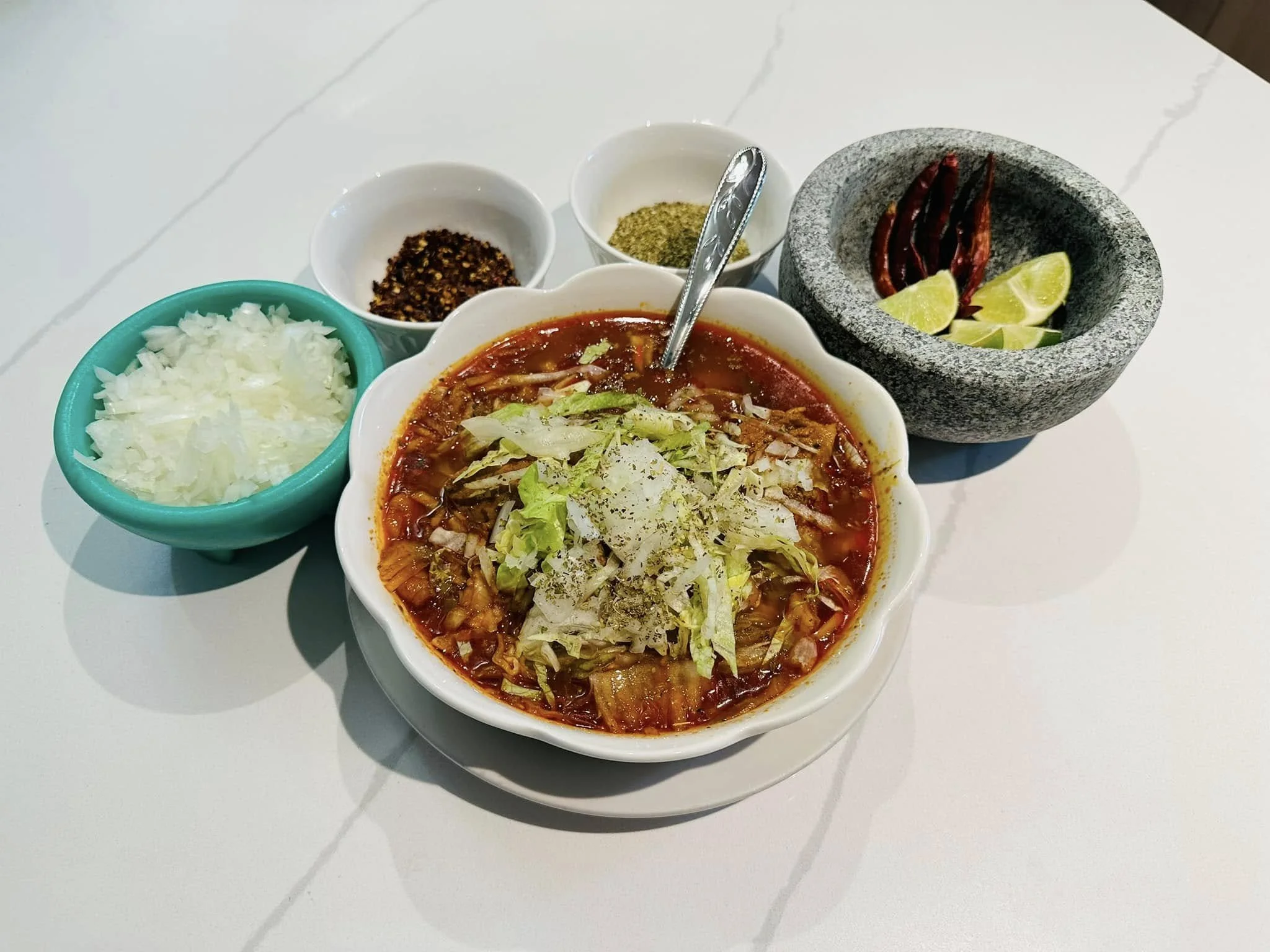 A bowl of Mexican red bean soup topped with shredded lettuce, served alongside diced white onions, dried chili peppers, lime wedges, and various spices in small bowls.