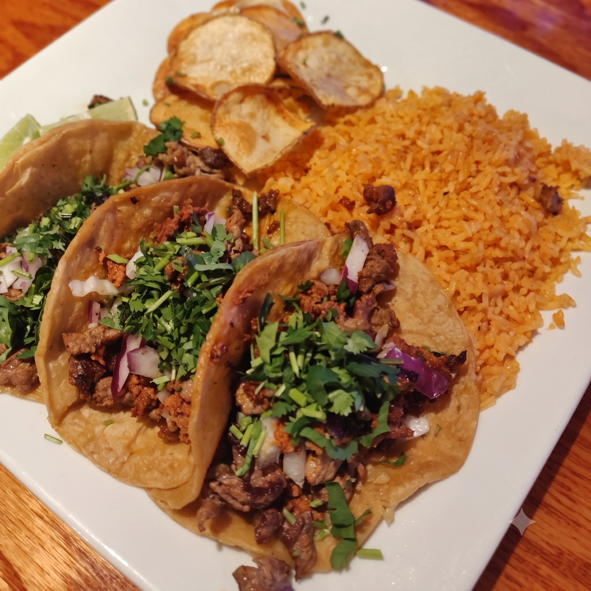 Several tacos filled with meat, chopped onions, and cilantro, served with a side of Mexican-style rice and potato chips on a white plate.