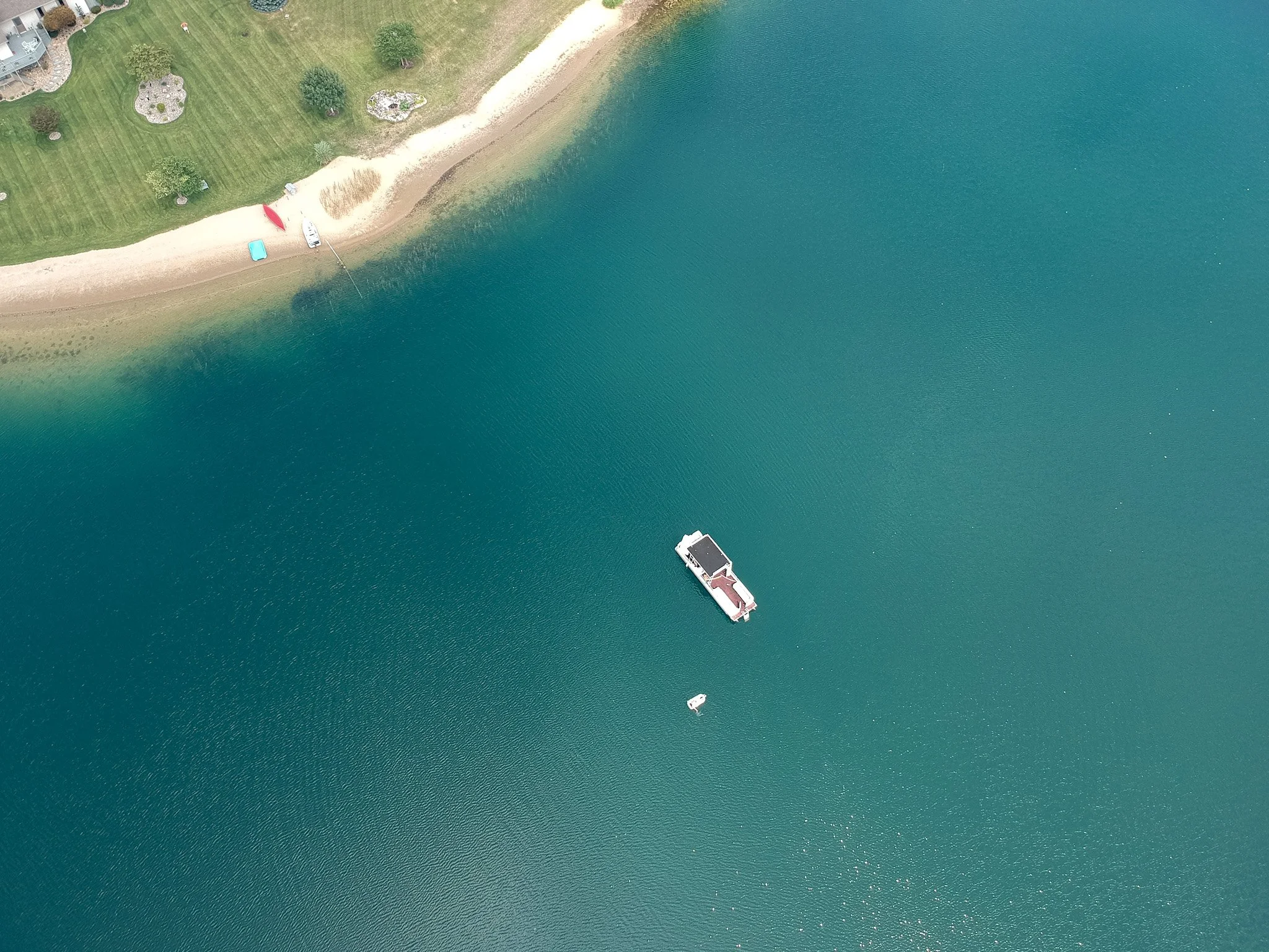 An aerial view of a waterfront property with a sandy beach, boat dock, and a grassy yard with trees, and a boat in the water.