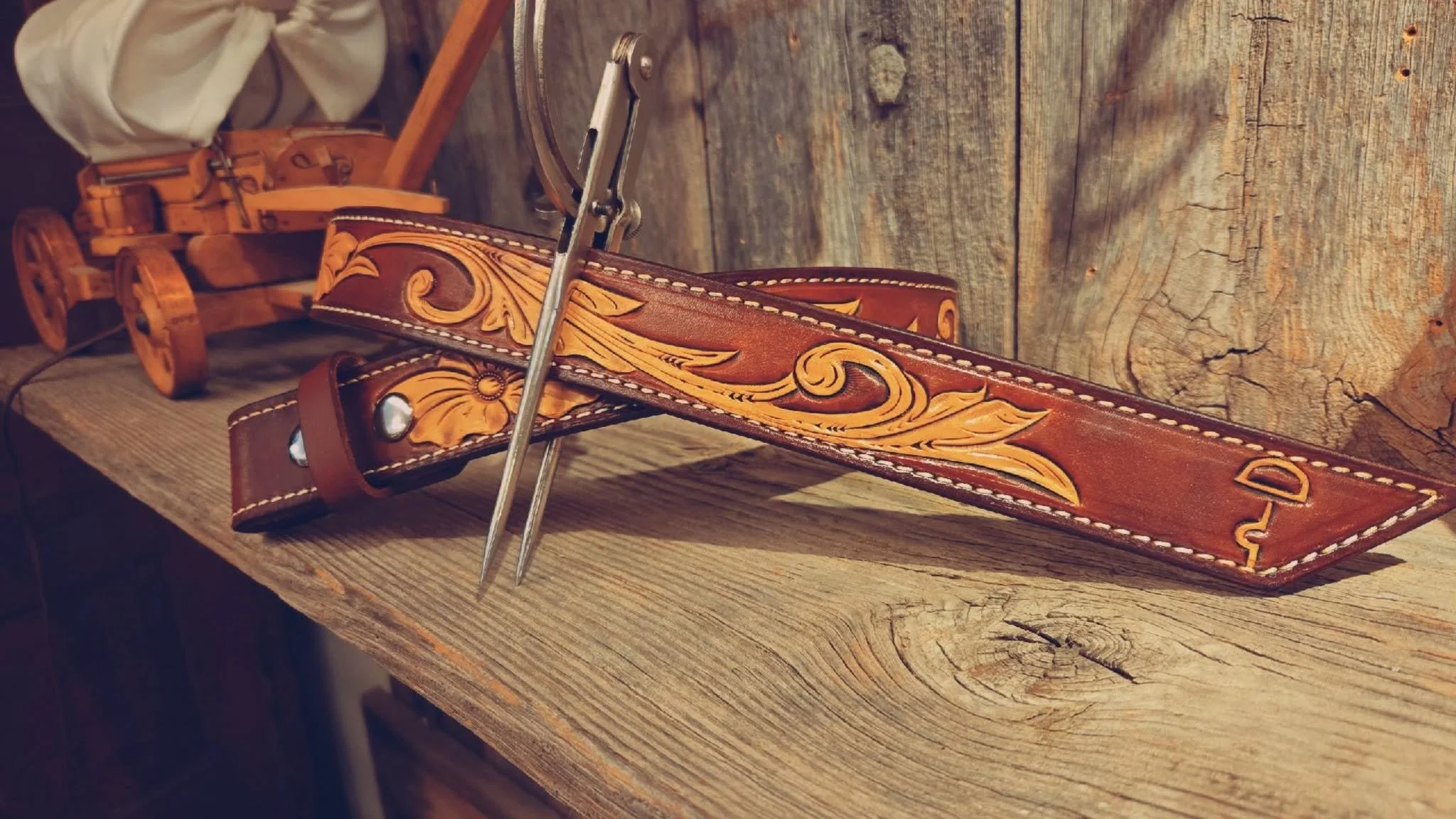 Leather belt with ornate embossed design resting on a wooden surface with leather and wooden toy train in the background.