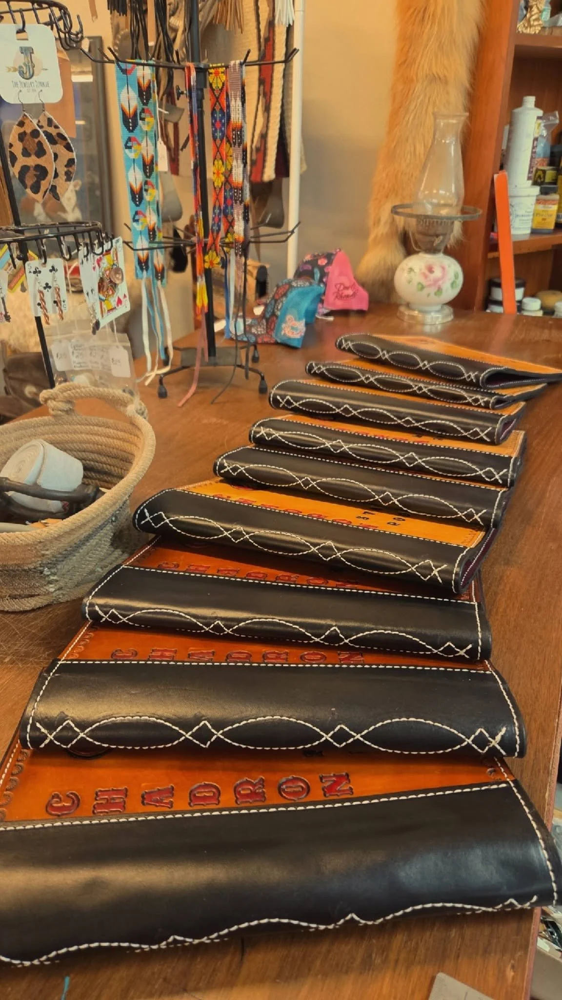 A display of black leather slots with white stitching, labeled with letters and numbers, on a wooden table. In the background, various jewelry pieces, including beaded necklaces and bracelets, are hanging on a black display stand. To the right, there