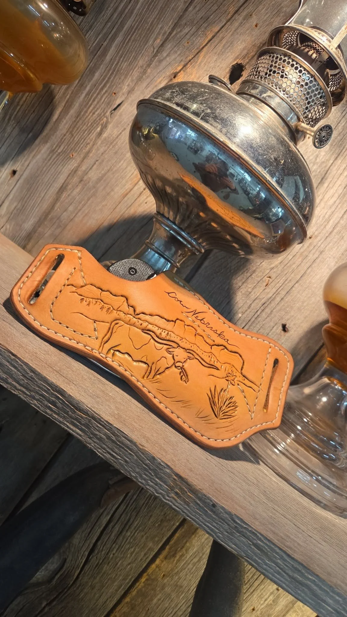 A vintage oil lantern, a leather house-shaped tray with mountain and desert scenes, and a glass of beer on a wooden table.