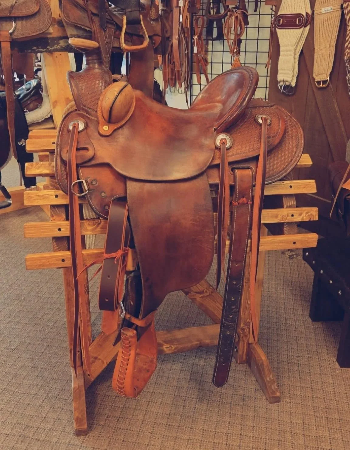 A leather Western-style saddle with stirrups, a saddle horn, and a set of leather straps, resting on a wooden saddle stand in a store with related riding gear hanging in the background.