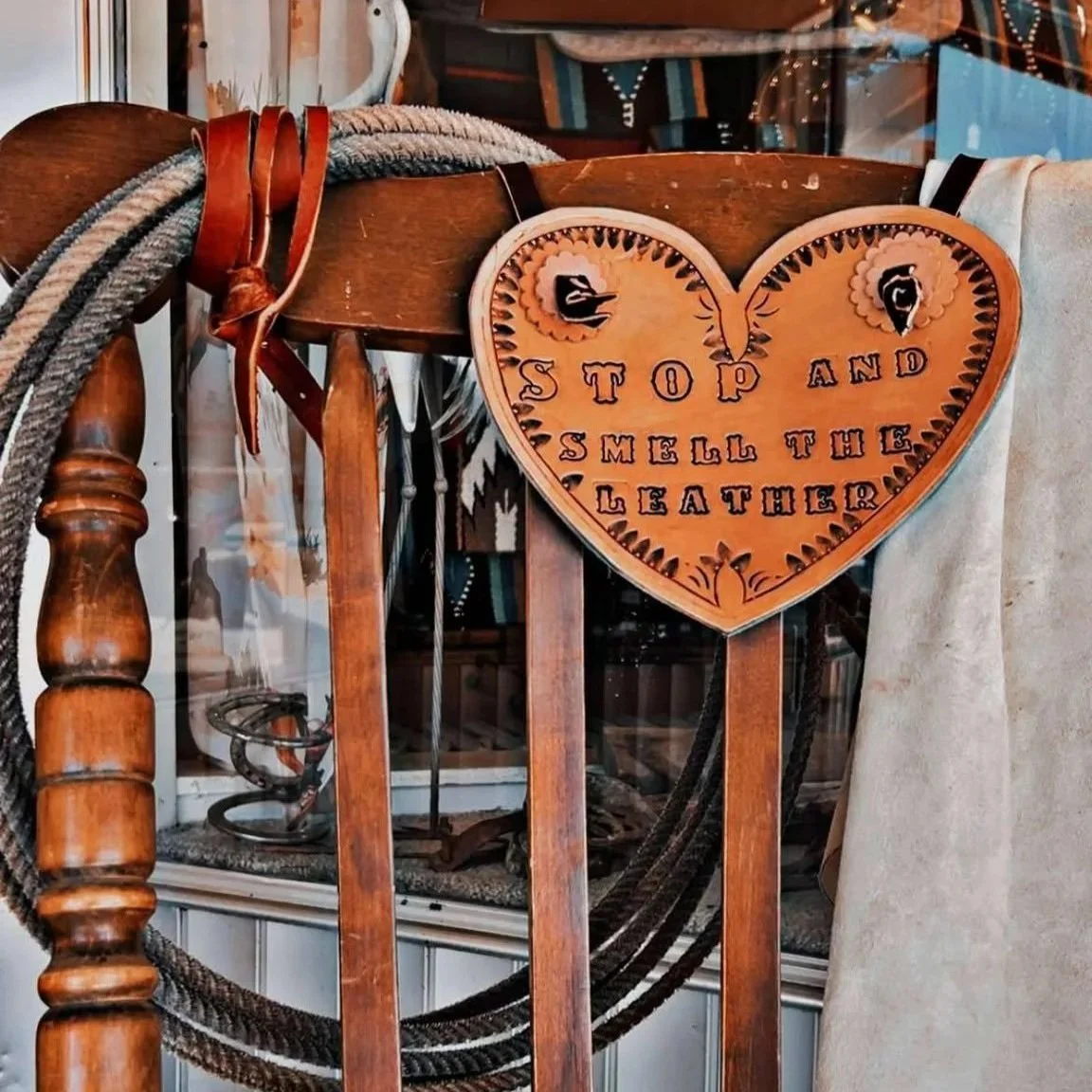 A wooden chair with a tied leather strap and a leather heart-shaped sign that reads "Stop and Smell the Leather" hanging on the back.