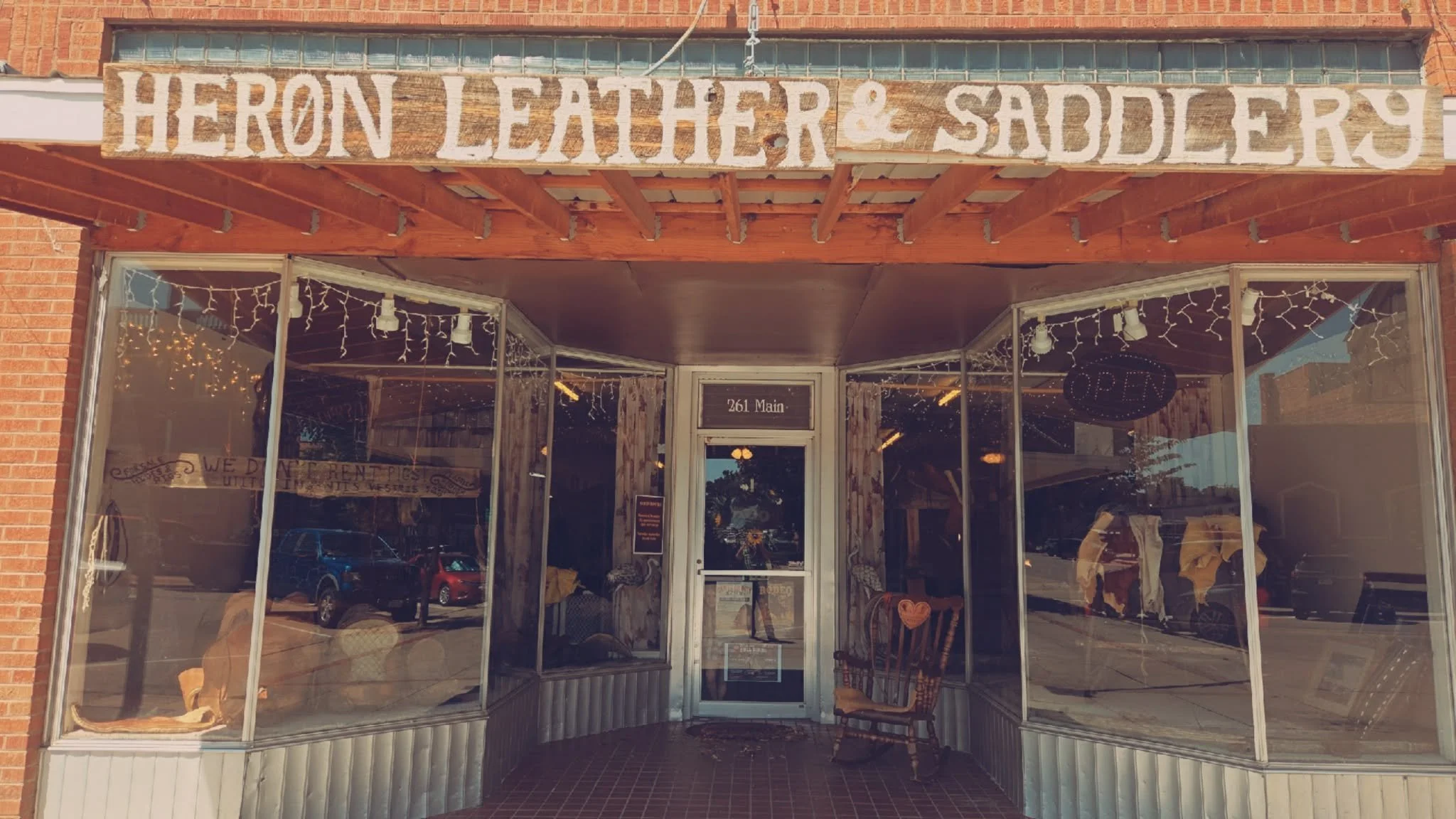 Storefront with a wooden sign 'HERON LEATHER & SADDLERY', large glass windows, string lights, and an 'OPEN' sign, located at 261 Main.
