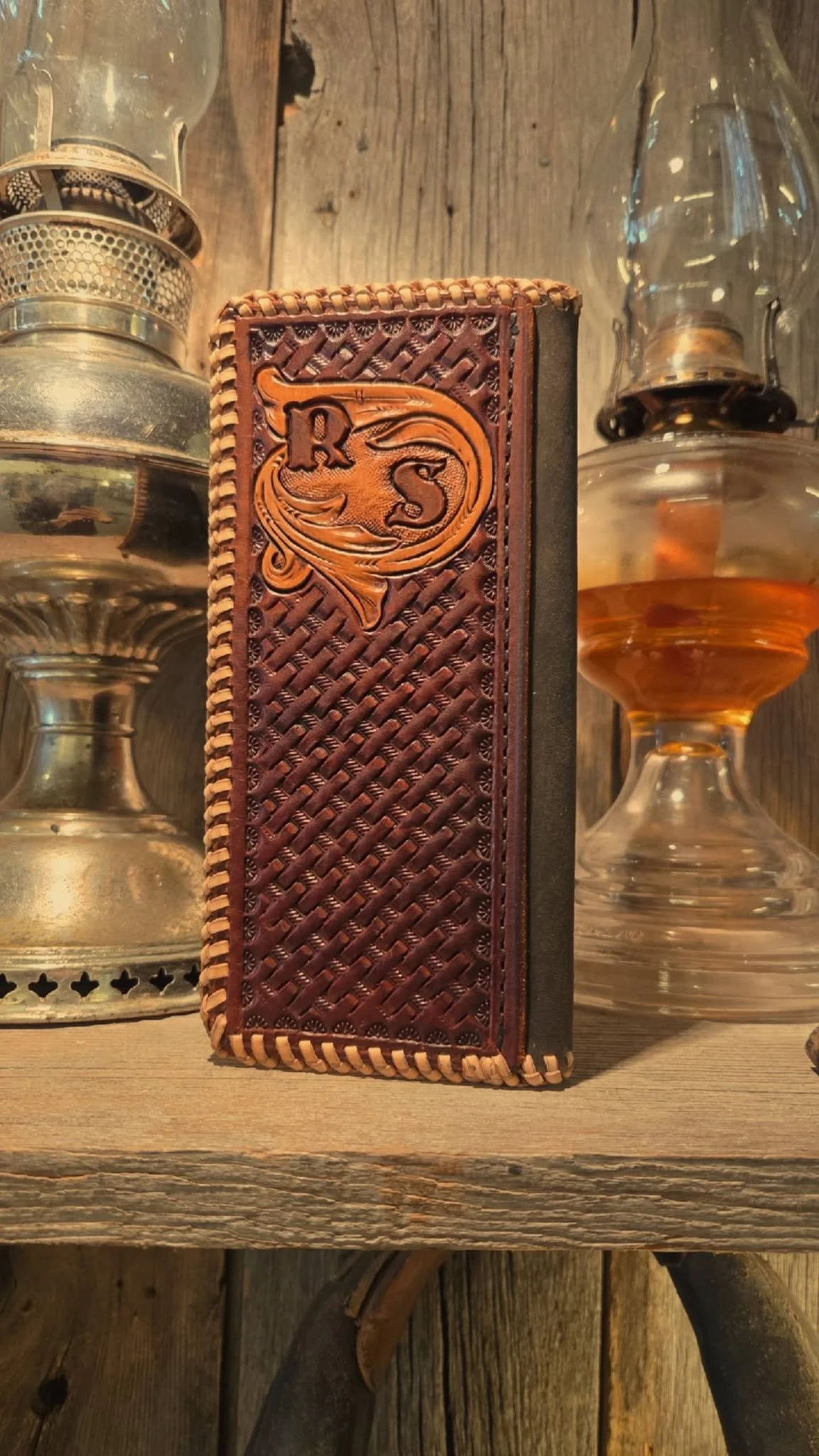A leather-bound book with a decorative embossed cover featuring a dragon and the initials 'R S' is positioned on a rustic wooden table, flanked by two vintage oil lanterns.