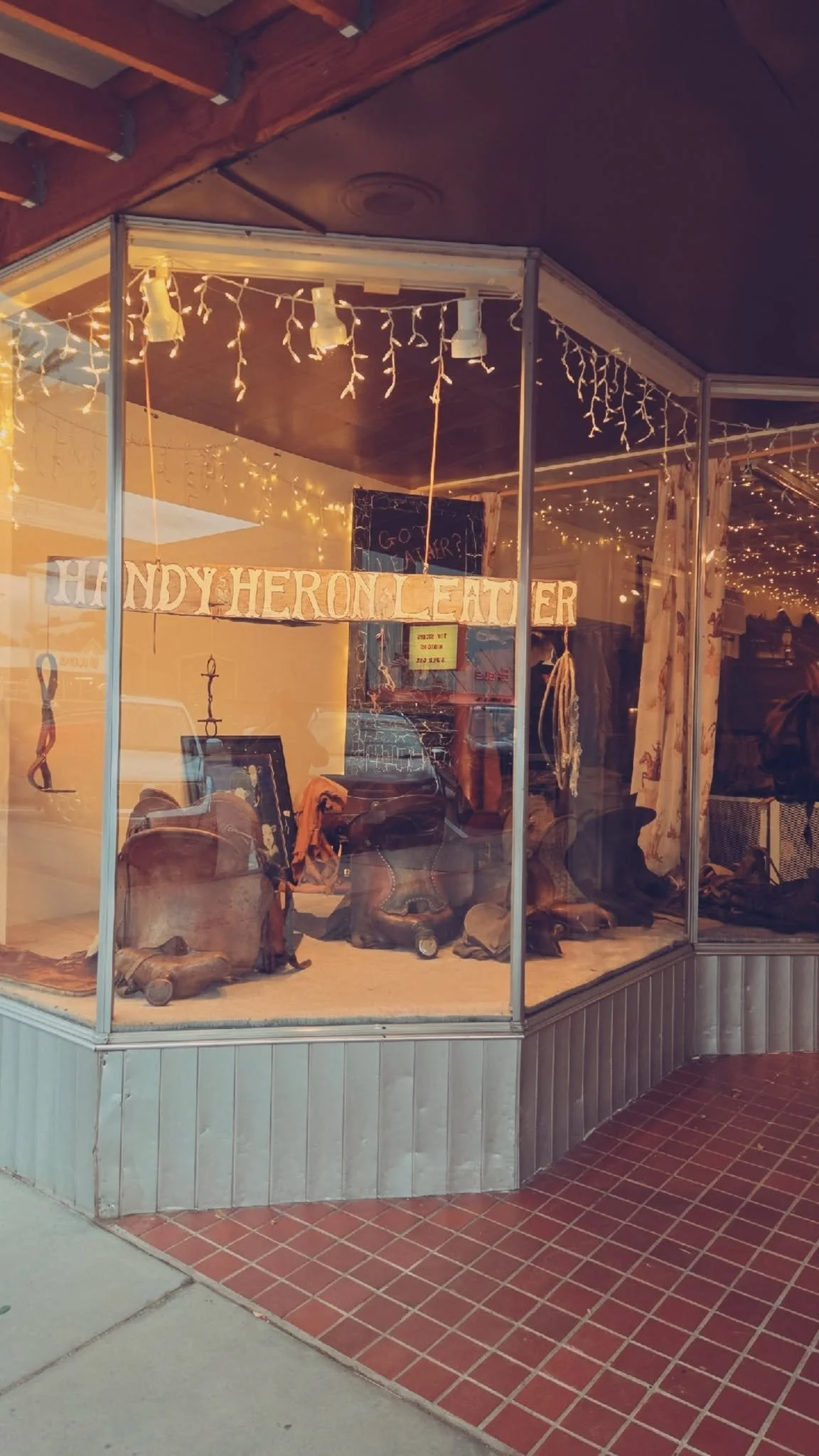 Storefront display of leather goods, including boots and slippers, with a sign that reads 'HANDY HERON CLEATHER' and hanging string lights inside.