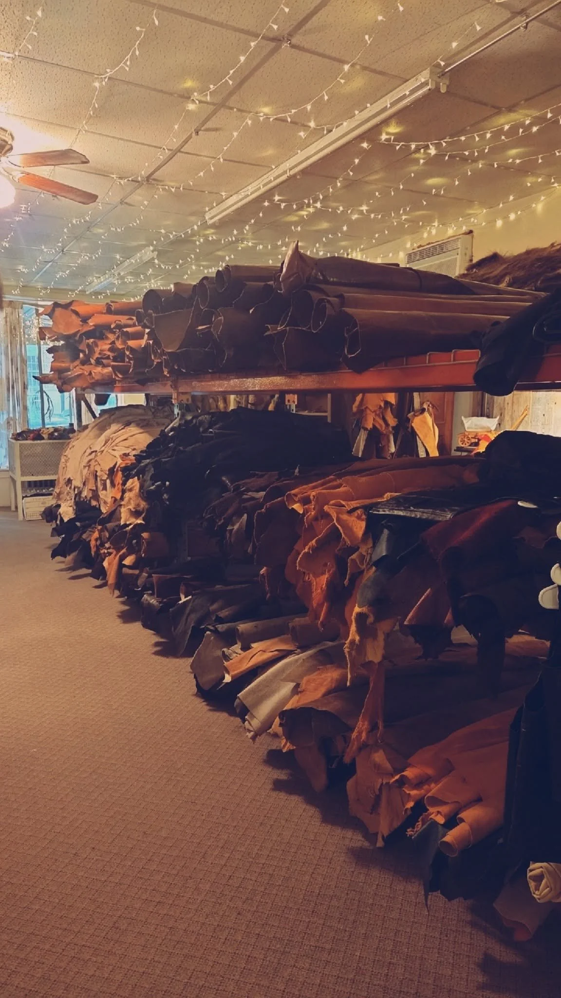 Interior view of a fabric or leather store with multiple shelves filled with rolls of material, and string lights hanging from the ceiling.