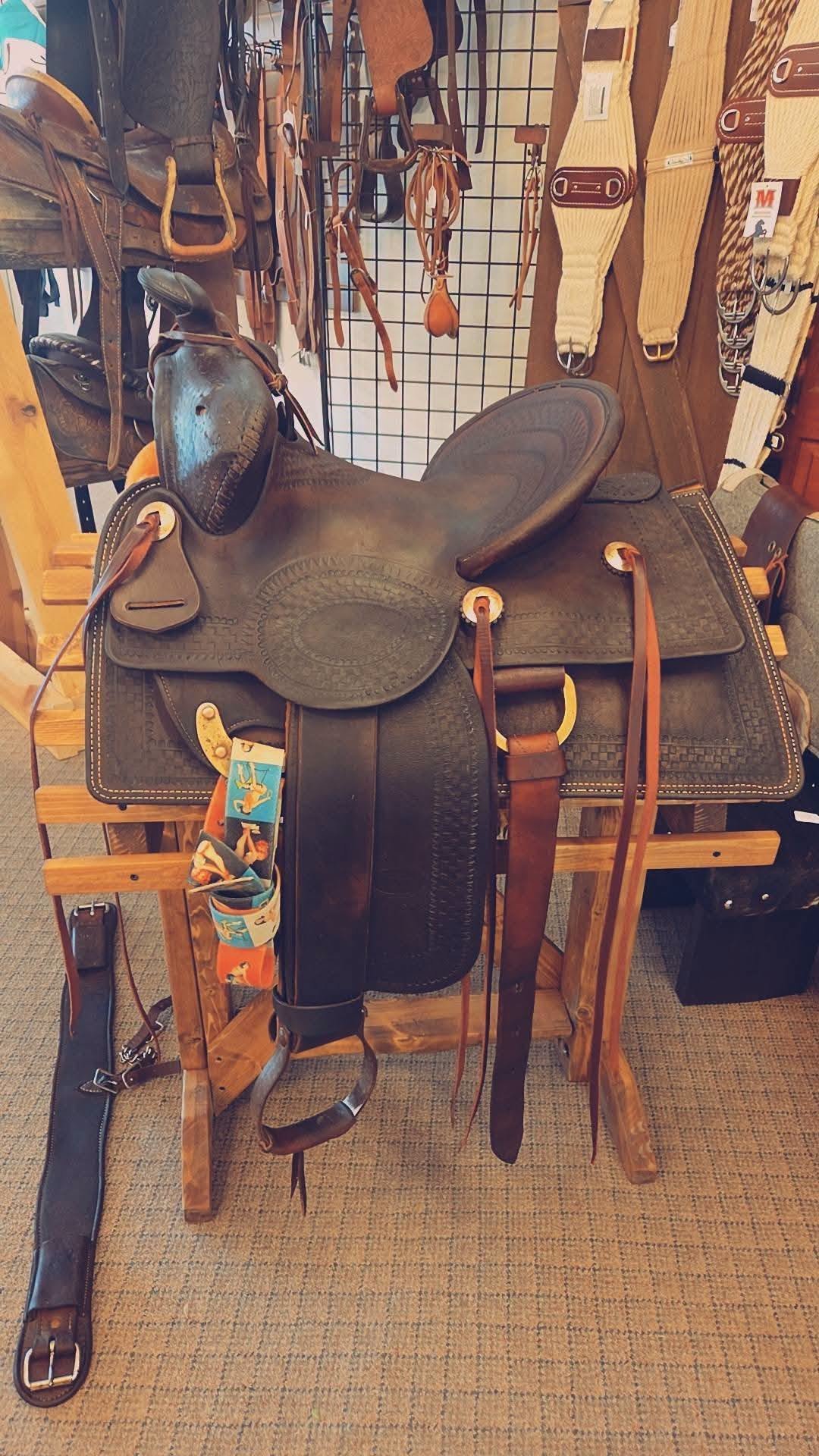 A leather western saddle on a wooden saddle stand surrounded by various horse tack and accessories displayed on a grid wall.