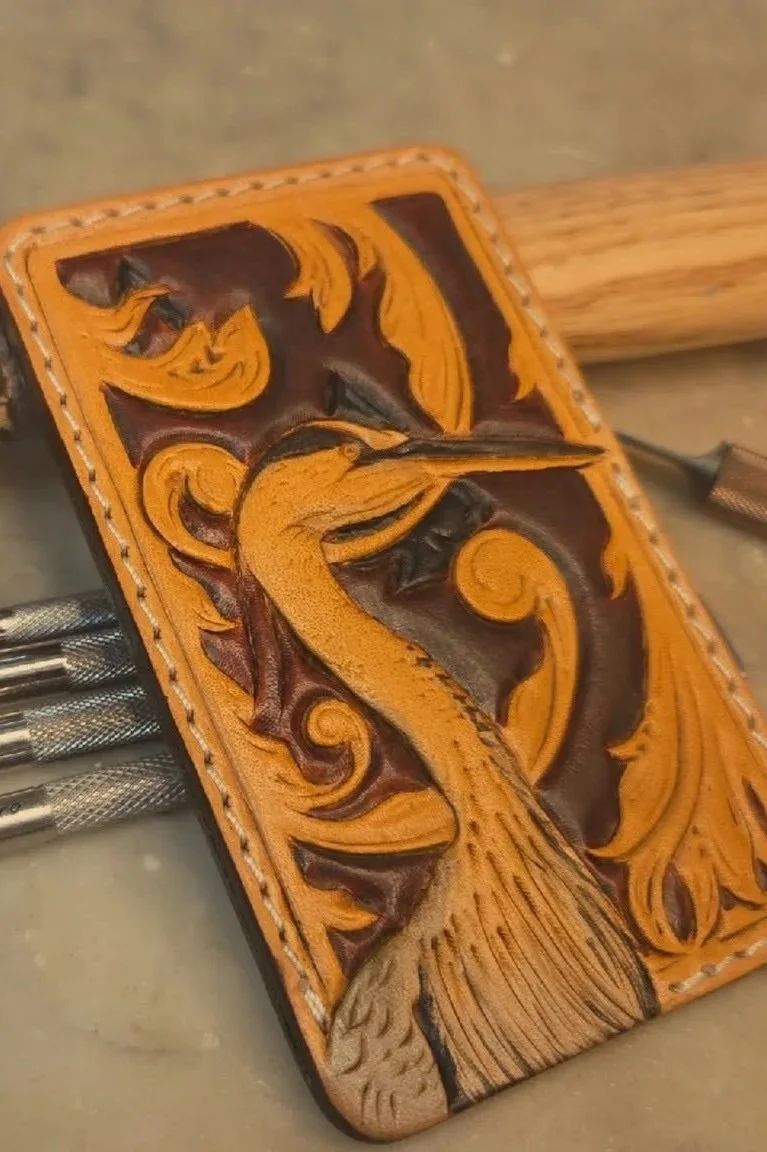 Close-up of a leather carving of a peacock with ornate scrolling patterns, attached to a wooden handle, resting on a work surface.