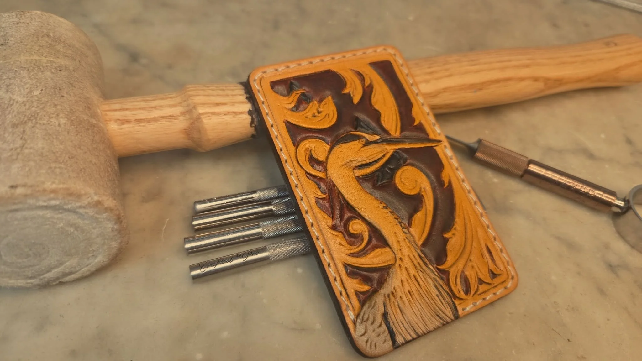 A leather keychain with intricate embossed design, various metal tools, and pairs of leather stamping and carving tools on a marble surface.
