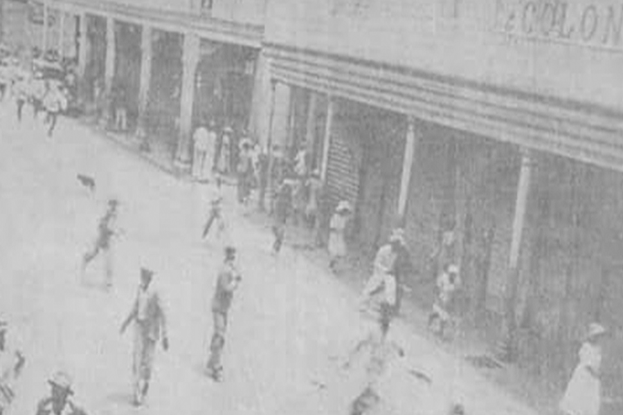 Historical black and white photo of people walking and standing on a city street with storefronts.