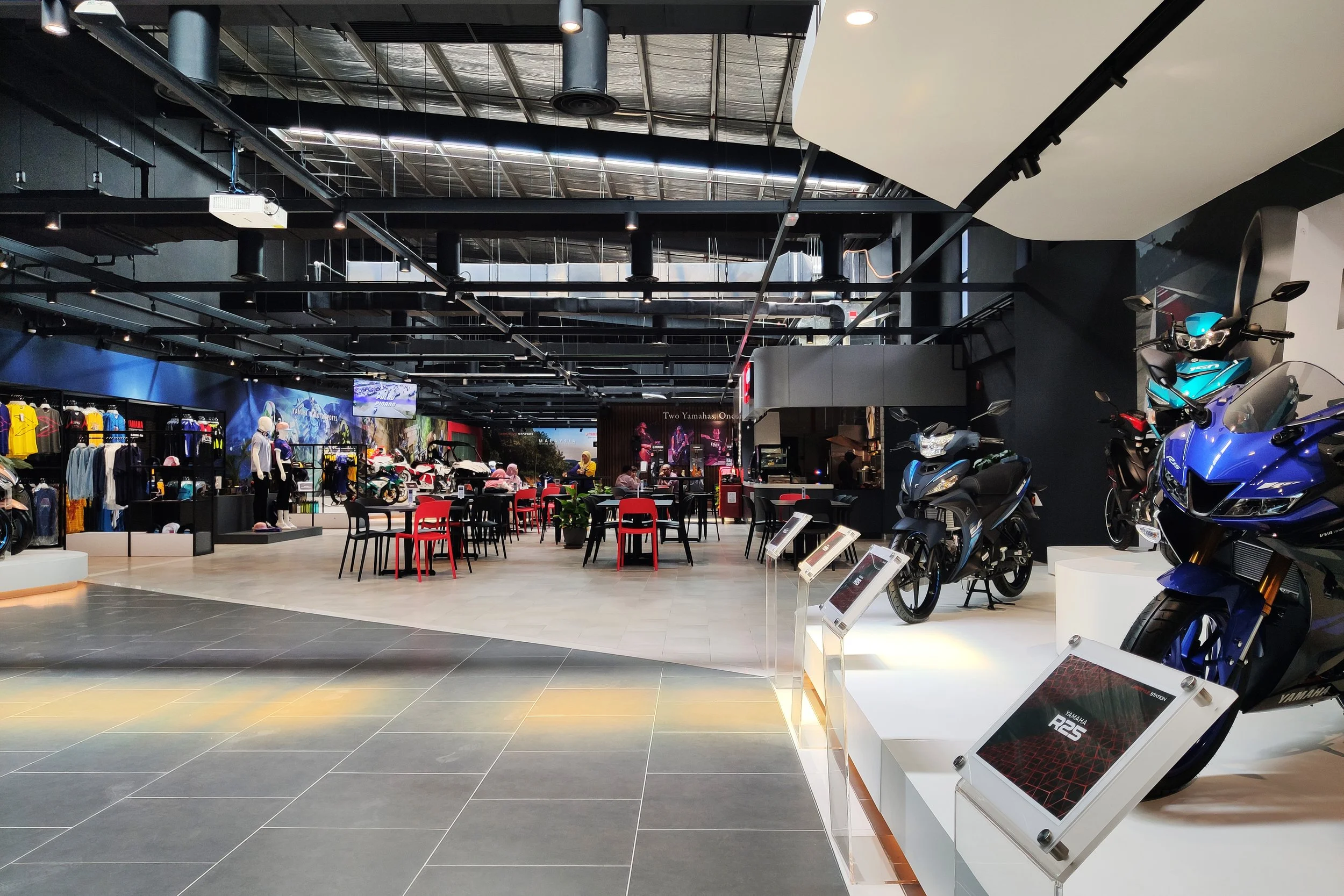 Interior of a modern motorcycle showroom featuring several motorcycles on display, apparel, mannequins, and a seating area with red and black chairs.