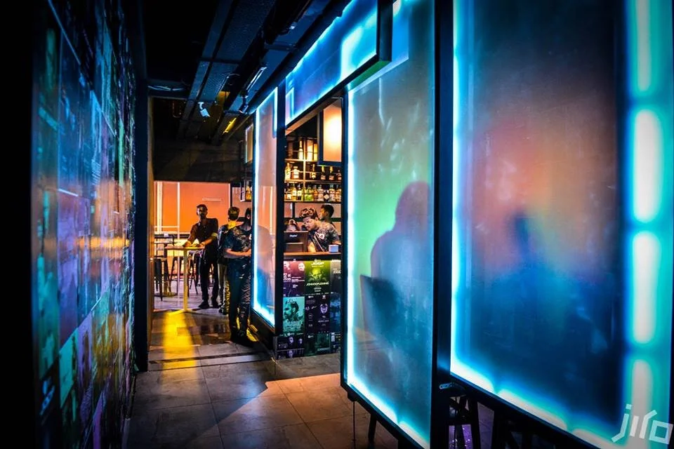 A futuristic bar interior with neon blue lighting, people standing near shelves of bottles, and colorful illuminated walls.