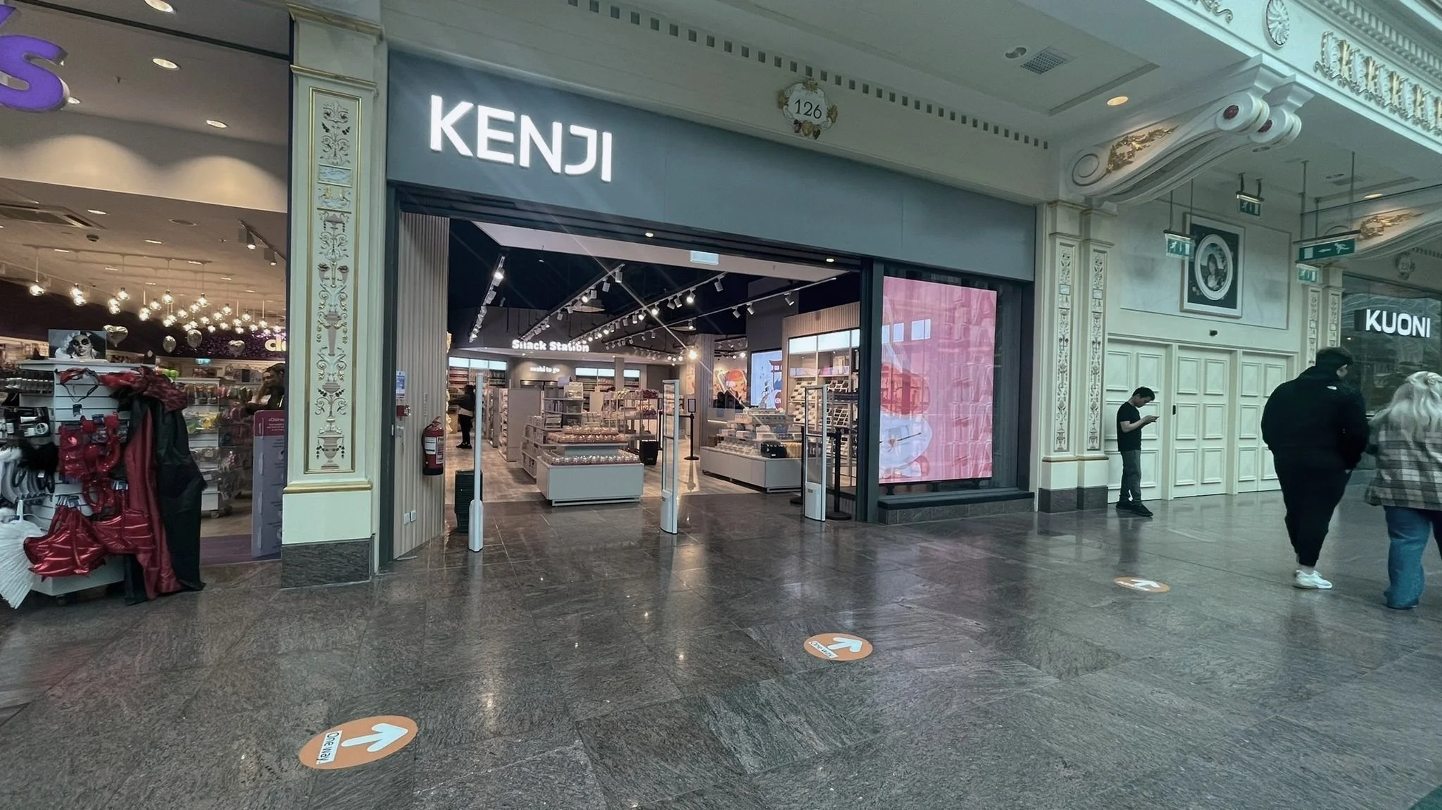 Entrance to Kenji store in a shopping mall, featuring a snack station display. Nearby stores and shoppers are visible in the mall corridor.