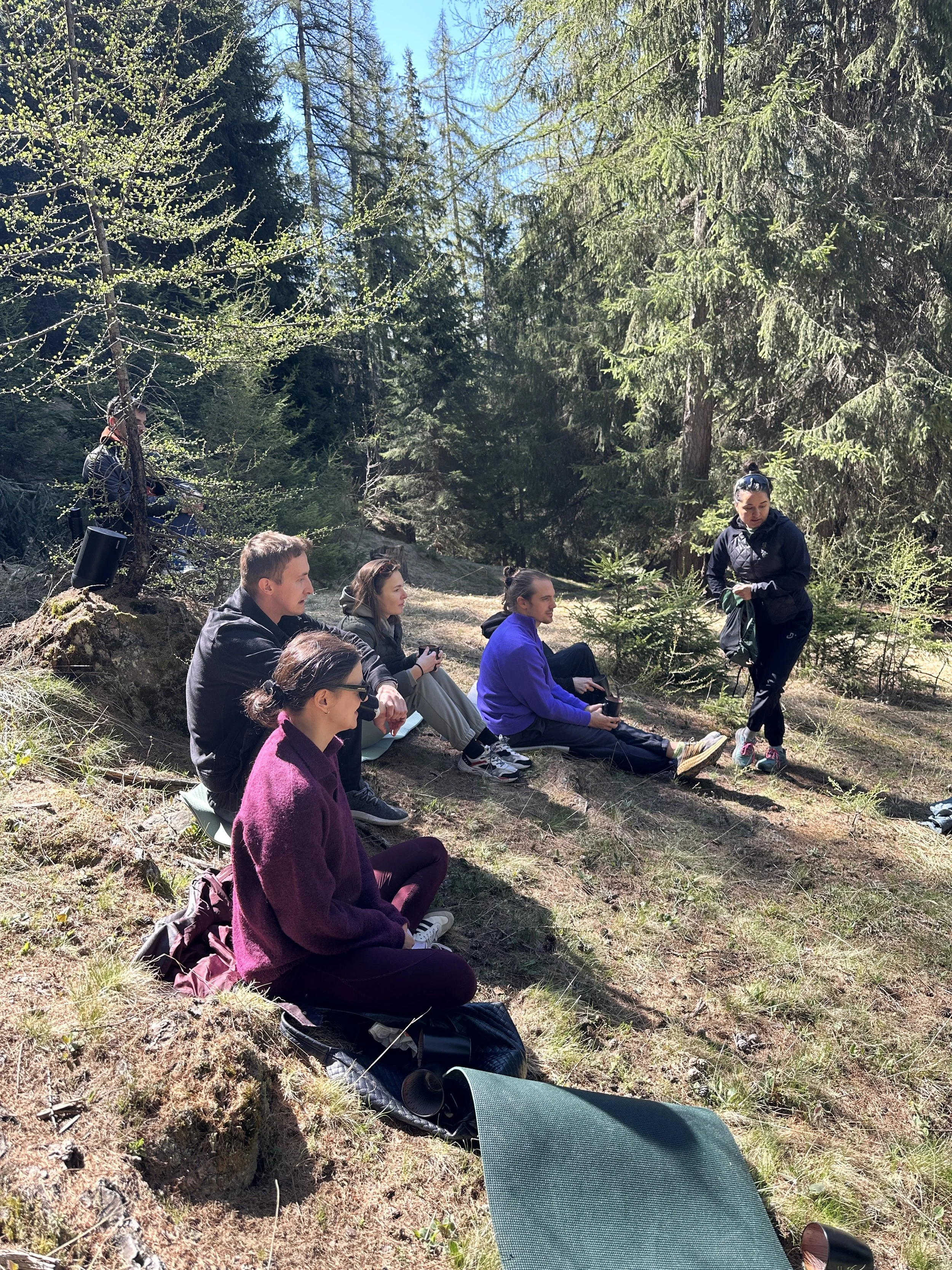 A group of people sitting on the ground in a forest, with a woman standing and talking nearby, surrounded by tall trees and green foliage on a sunny day.
