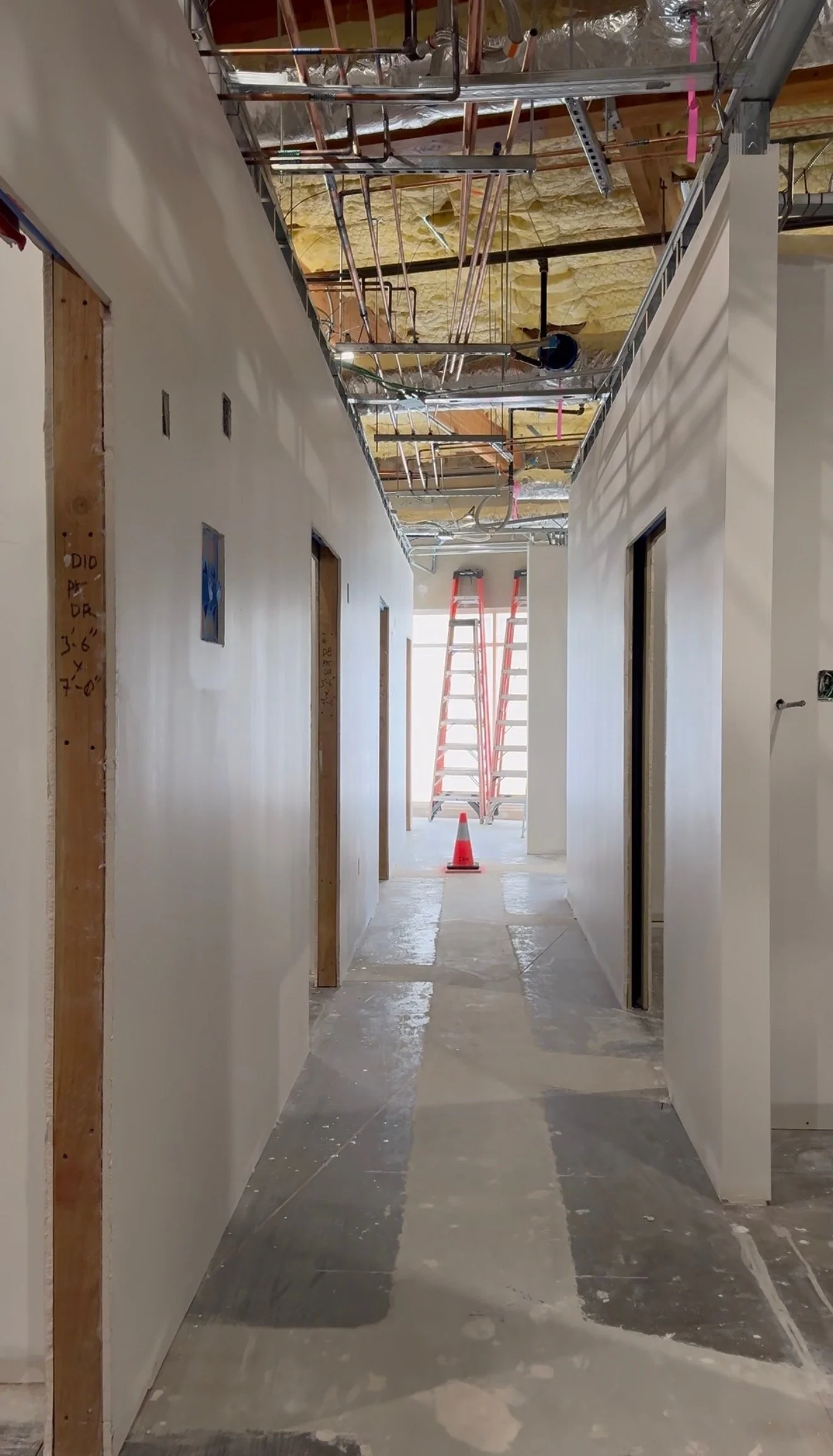 Construction corridor with exposed ceiling pipes, insulation, wall frames, ladder, orange cone, and sunlight, managed through construction administration by Hesse Design & Build.