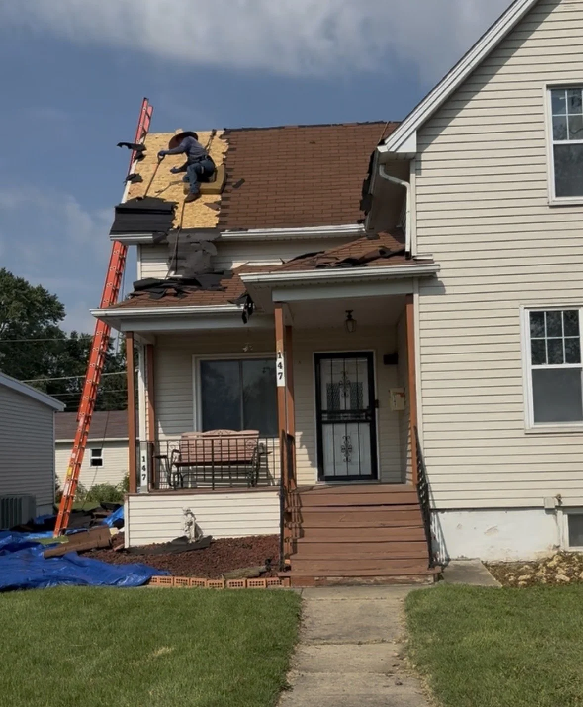 A house with a damaged roof undergoing repair. A worker is on the roof, replacing shingles, with a ladder leaning against the house.