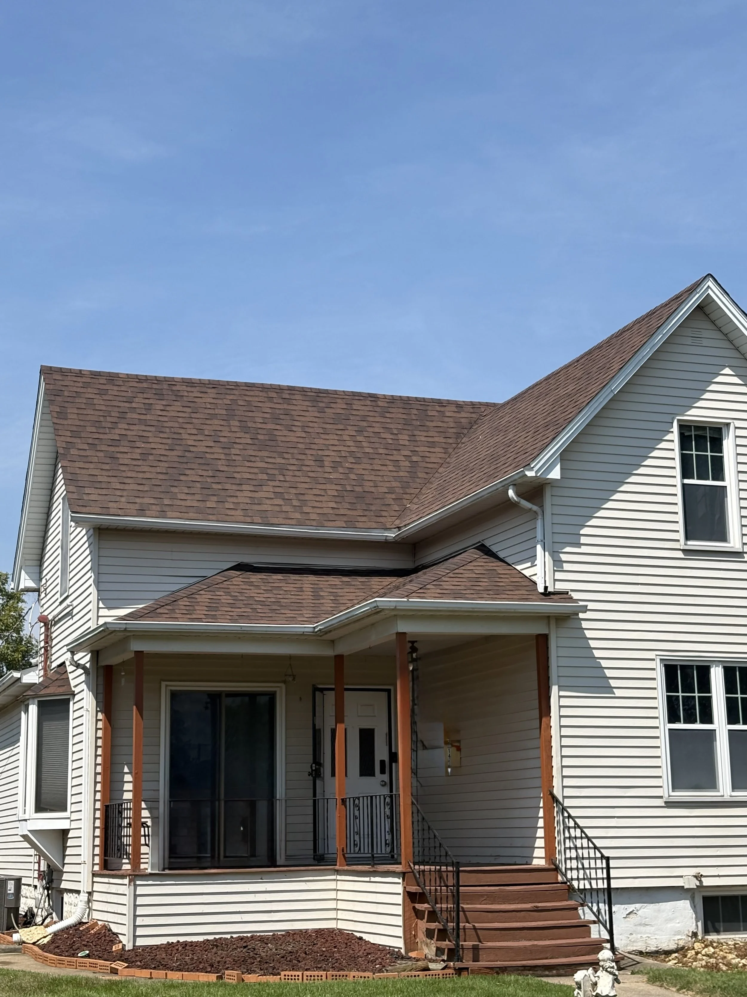 A house with light-colored siding and a red shingled roof under a blue sky. The backyard features a screened-in porch with stairs leading down to a freshly landscaped area with dark mulch and a small brick border.