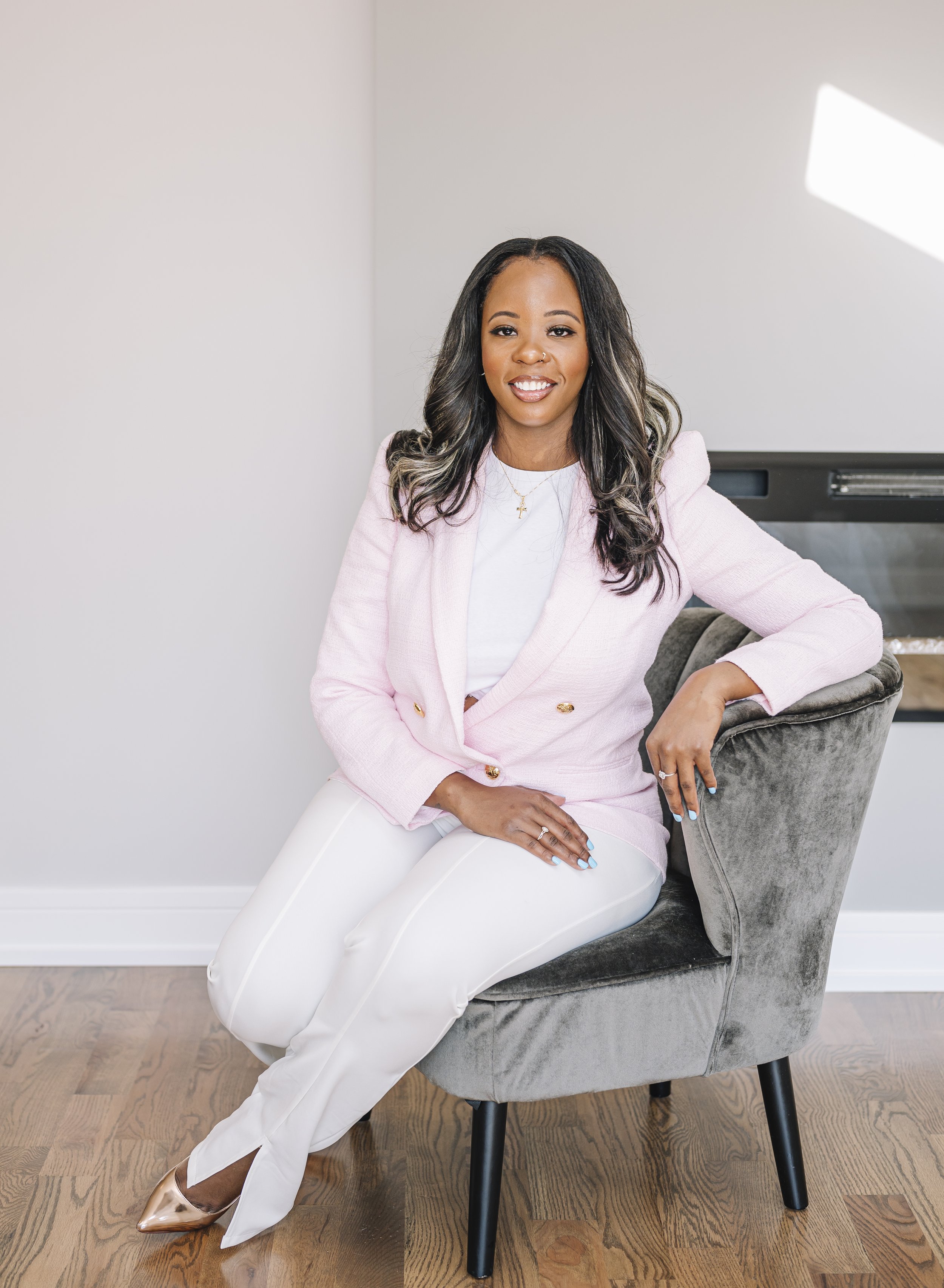 A woman with long wavy black hair and light brown skin sitting on a gray velvet chair in a bright room. She is wearing a pink blazer, white pants, and rose gold pointed-toe heels, with a gold necklace and rings, smiling at the camera.