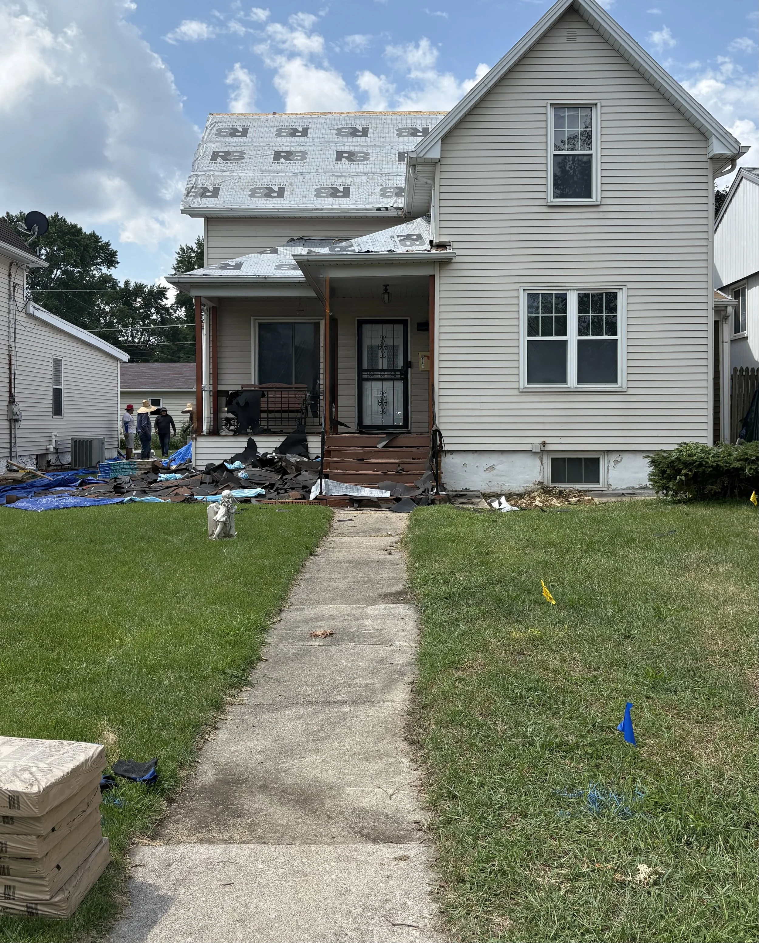 A house undergoing roof repair, with new roofing materials on the ground and the roof partially covered with roofing underlayment. A sidewalk runs from the front of the house into the yard, with a lawn on each side. Several workers are visible near the house, and the sky is partly cloudy.