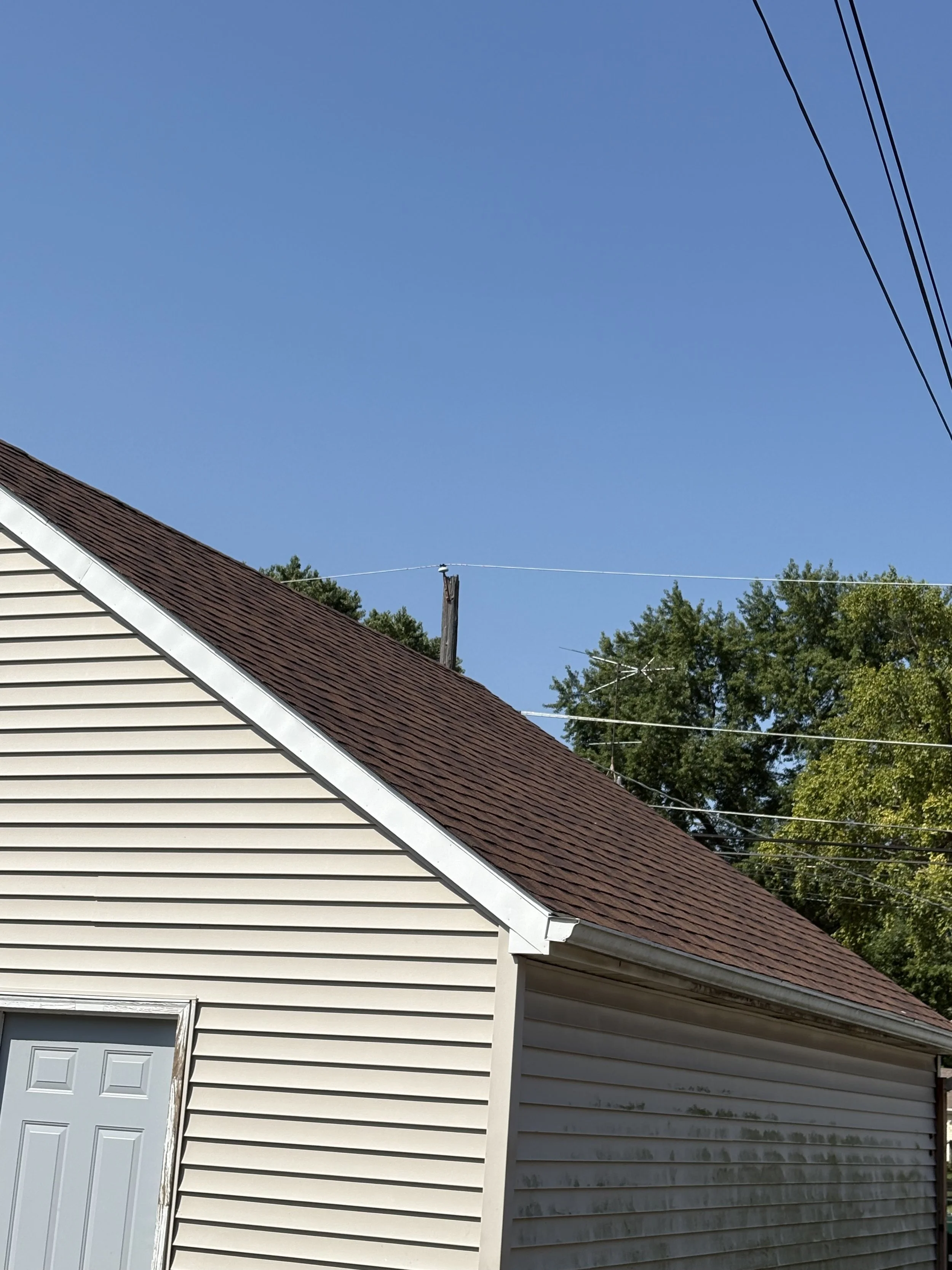 A house with beige siding and a brown shingled roof, a white garage door, a sloping roof edge, trees, and utility lines in the background under a clear blue sky.