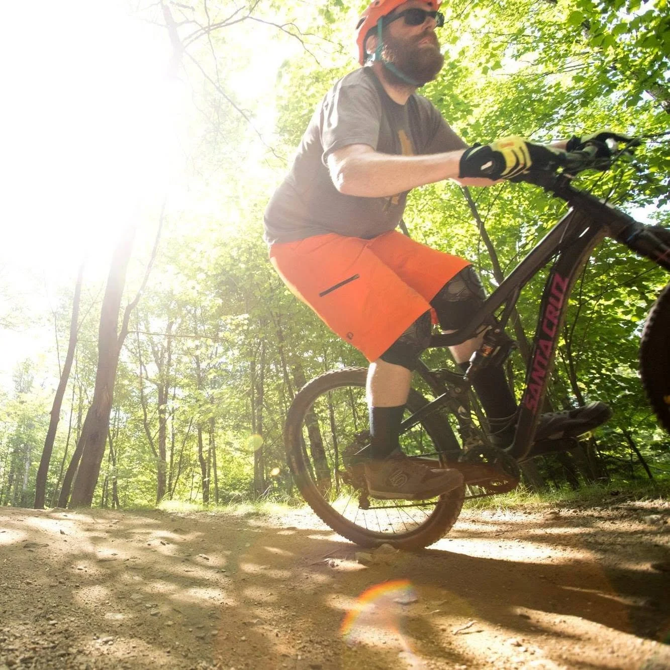 A man riding a mountain bike on a dirt trail in a wooded area with sunlight filtering through the trees.