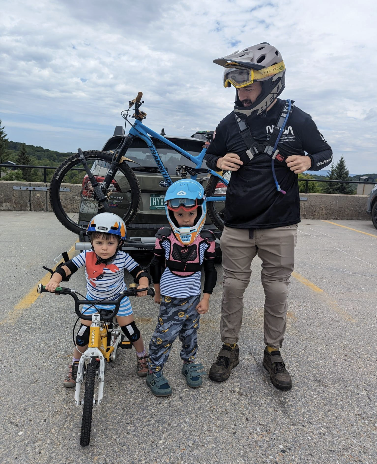 Three children with helmets and riding gear, posing with a man in a parking lot, with a mountain bike and mountain scenery in the background.