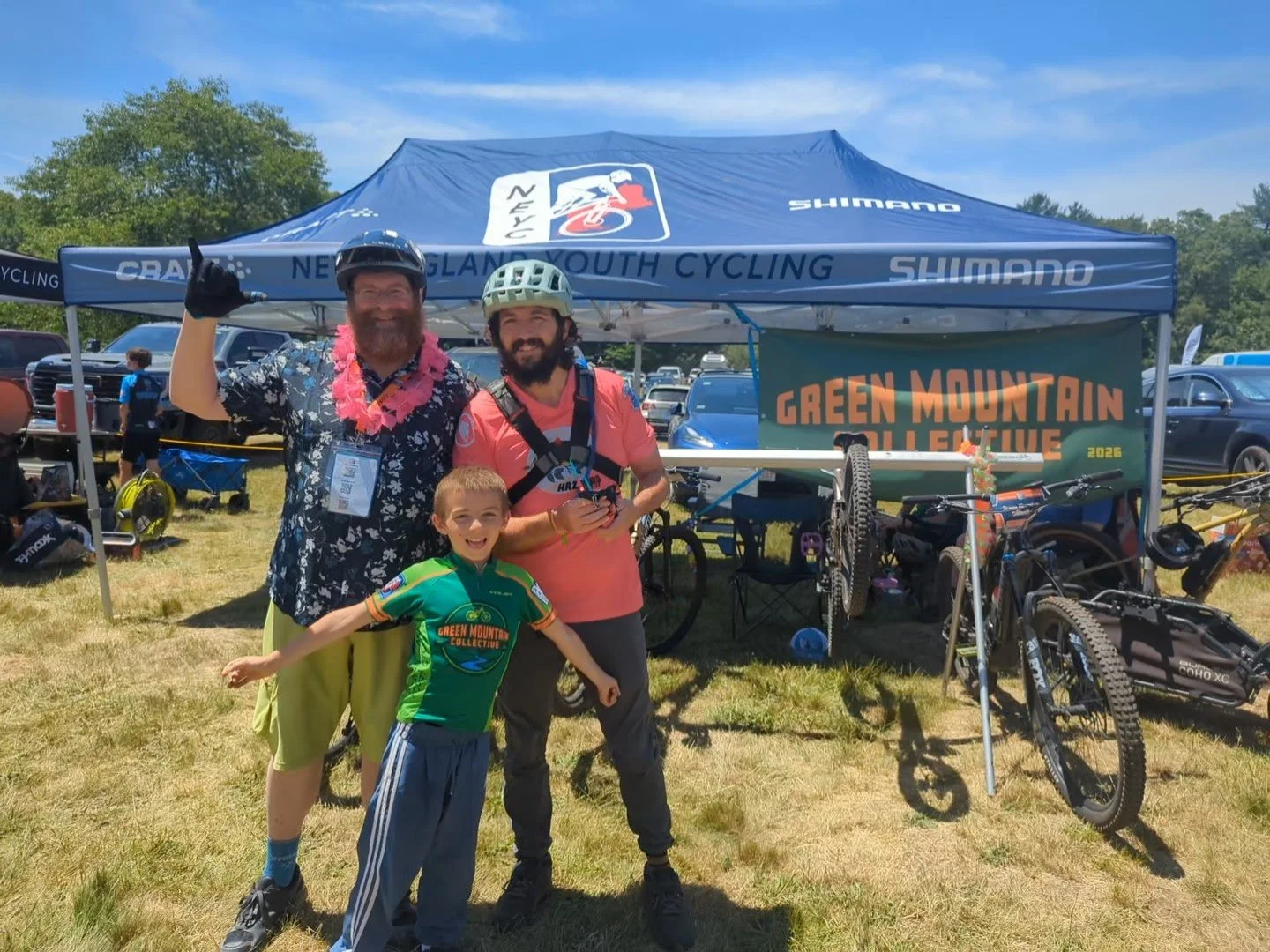 Three people, two adults and a child, standing in front of a cycling event tent at Green Mountain Bicycle Collective. The man on the left is wearing a colorful Hawaiian shirt, shorts, and a lei, pointing upward. The man on the right is wearing a helmet and pink shirt, holding a small item. The child in the front is wearing a green cycling jersey and gray pants, expanding his arms. The tent has a sign that reads 'Green Mountain Bicycle Collective' and features bicycles and cycling gear.