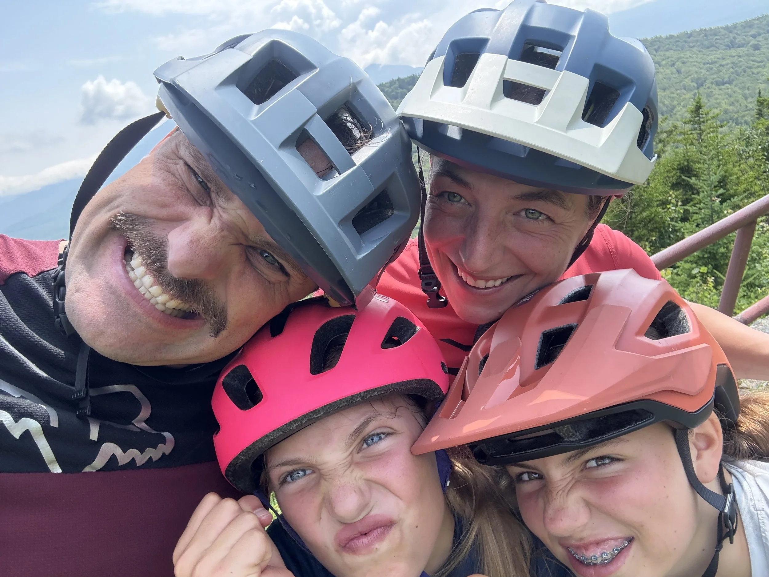 Group of five smiling people with helmets taking a selfie outdoors with mountains and blue sky in the background.
