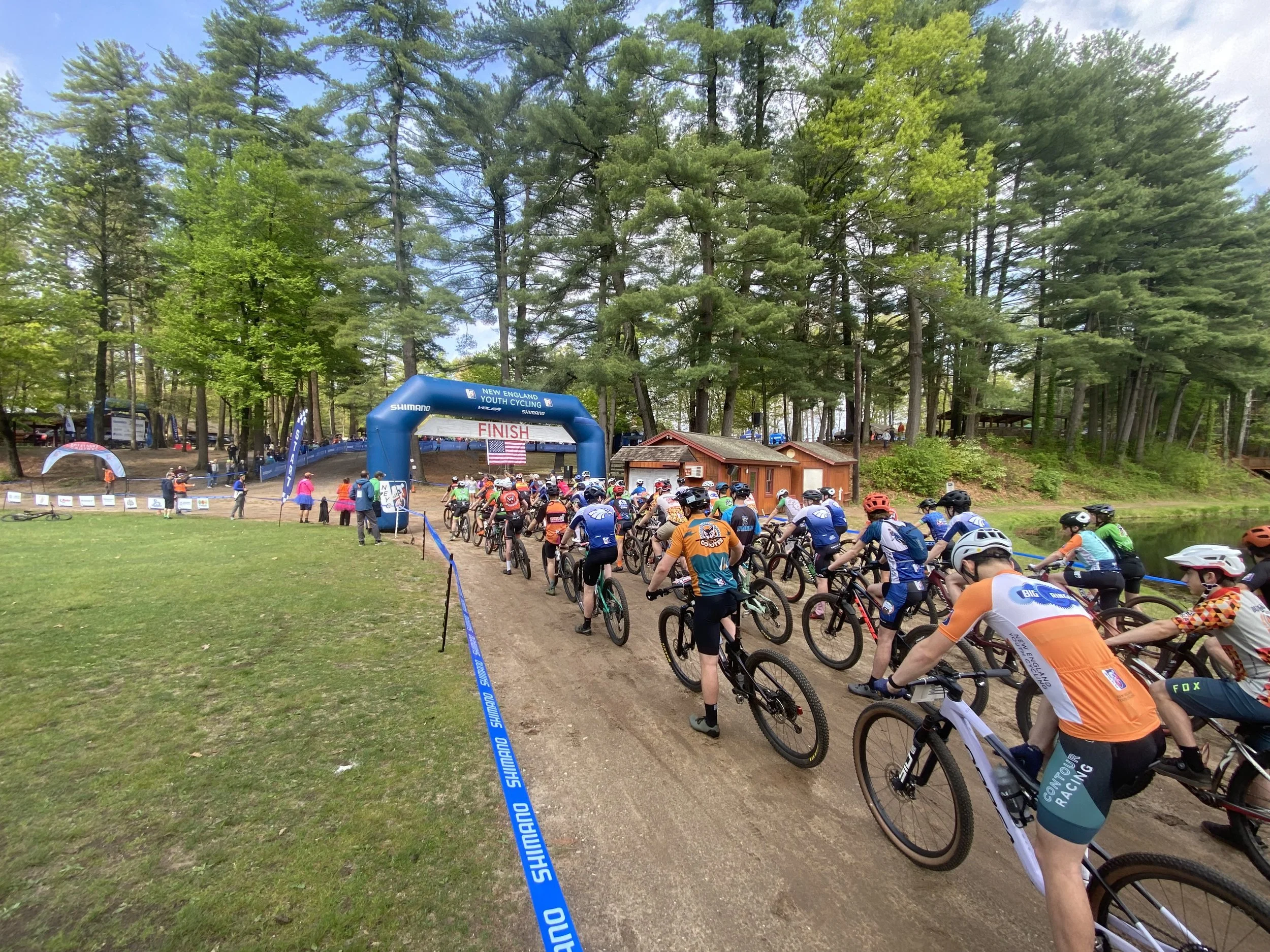 A large group of cyclists riding on a dirt trail through a wooded area near a finish line banner for a youth cycling race. The scene is shaded by tall green trees with a cloudy sky above.