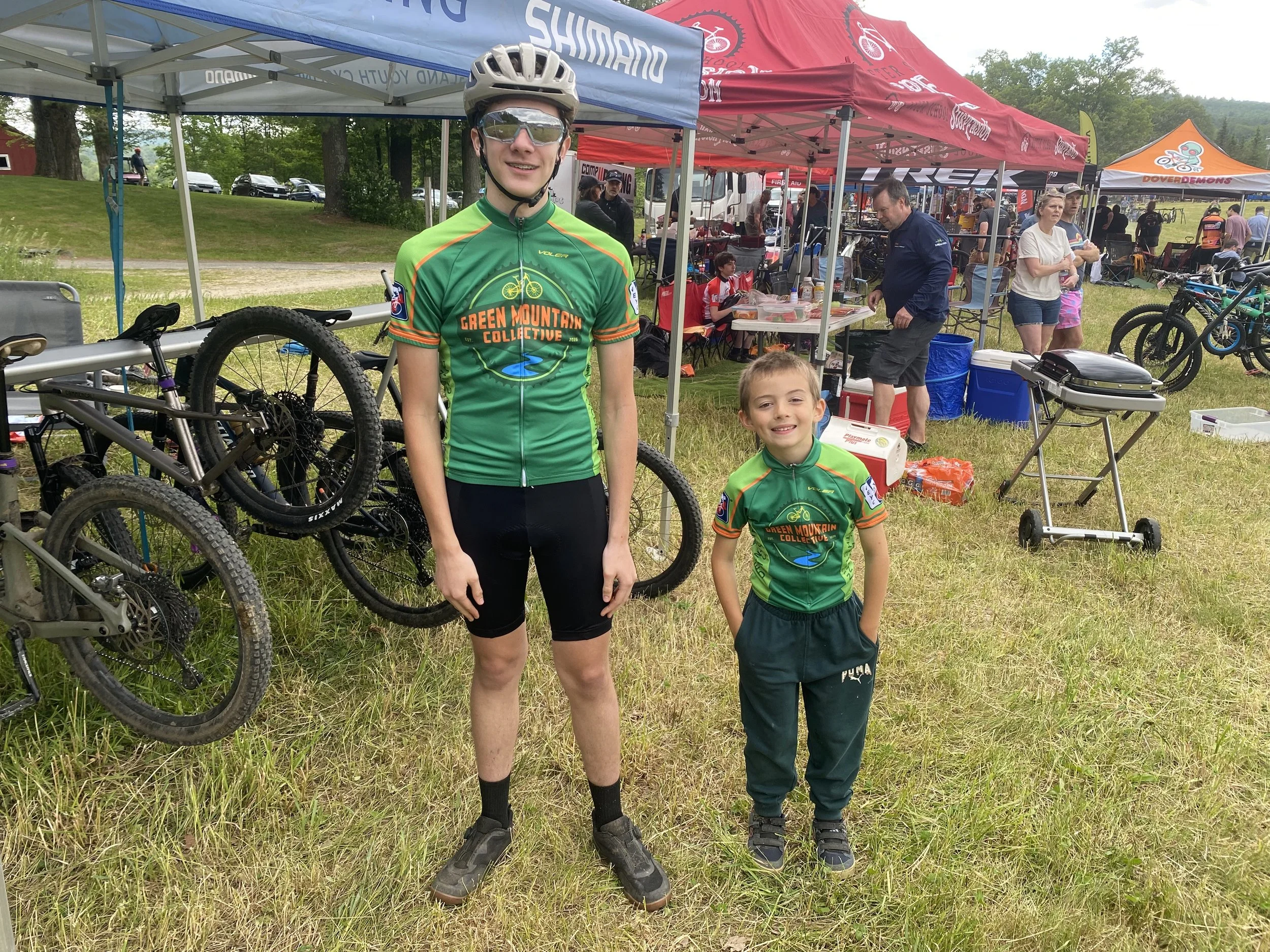 Two young boys in green cycling jerseys standing outdoors at a cycling event, with tents and other people in the background.