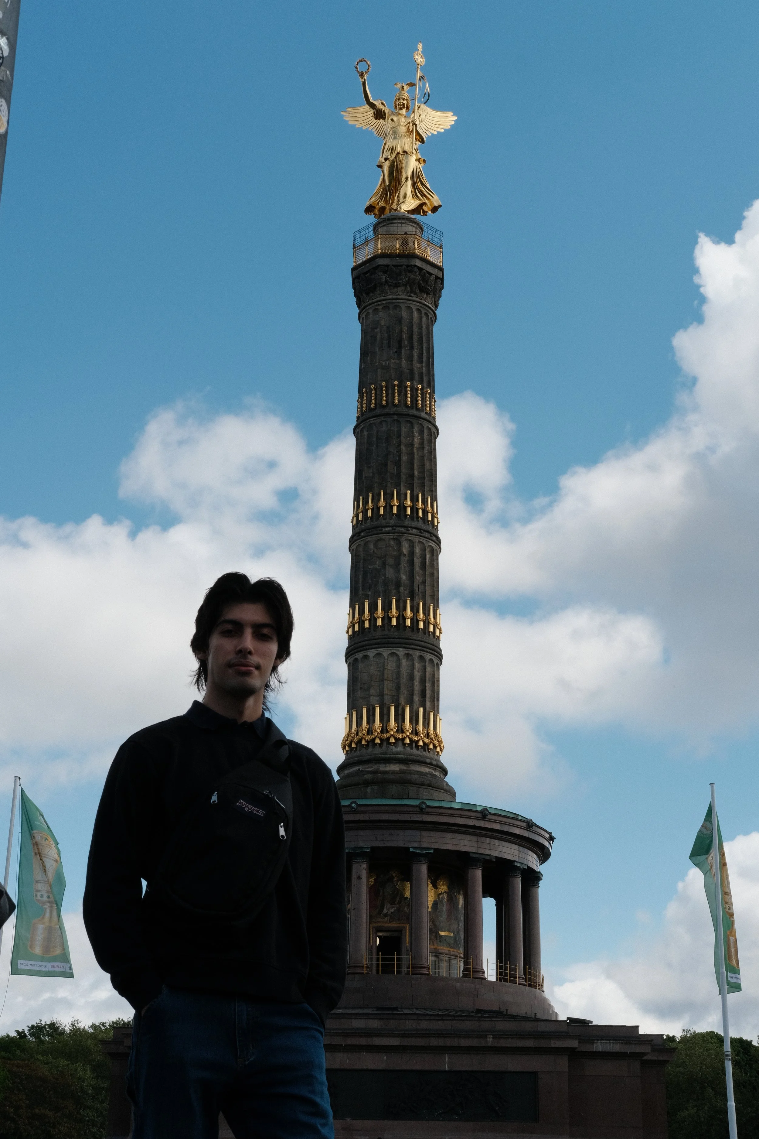 A young man with dark hair and a black jacket standing in front of the Victory Column in Berlin, Germany, with a partly cloudy sky in the background.