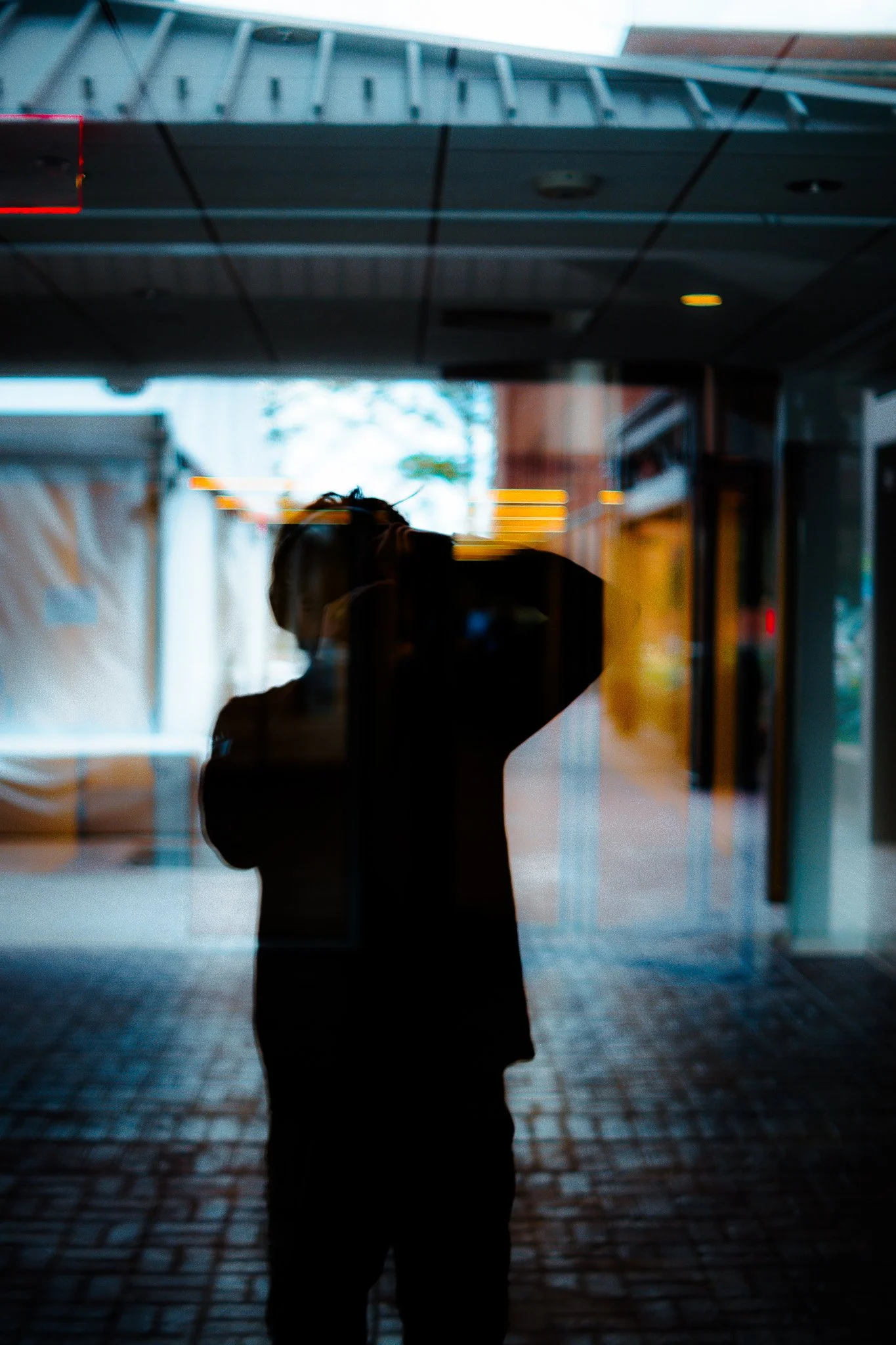 A silhouette of a young person standing outside near a glass window or door, with a blurred cityscape and building reflections visible in the background.