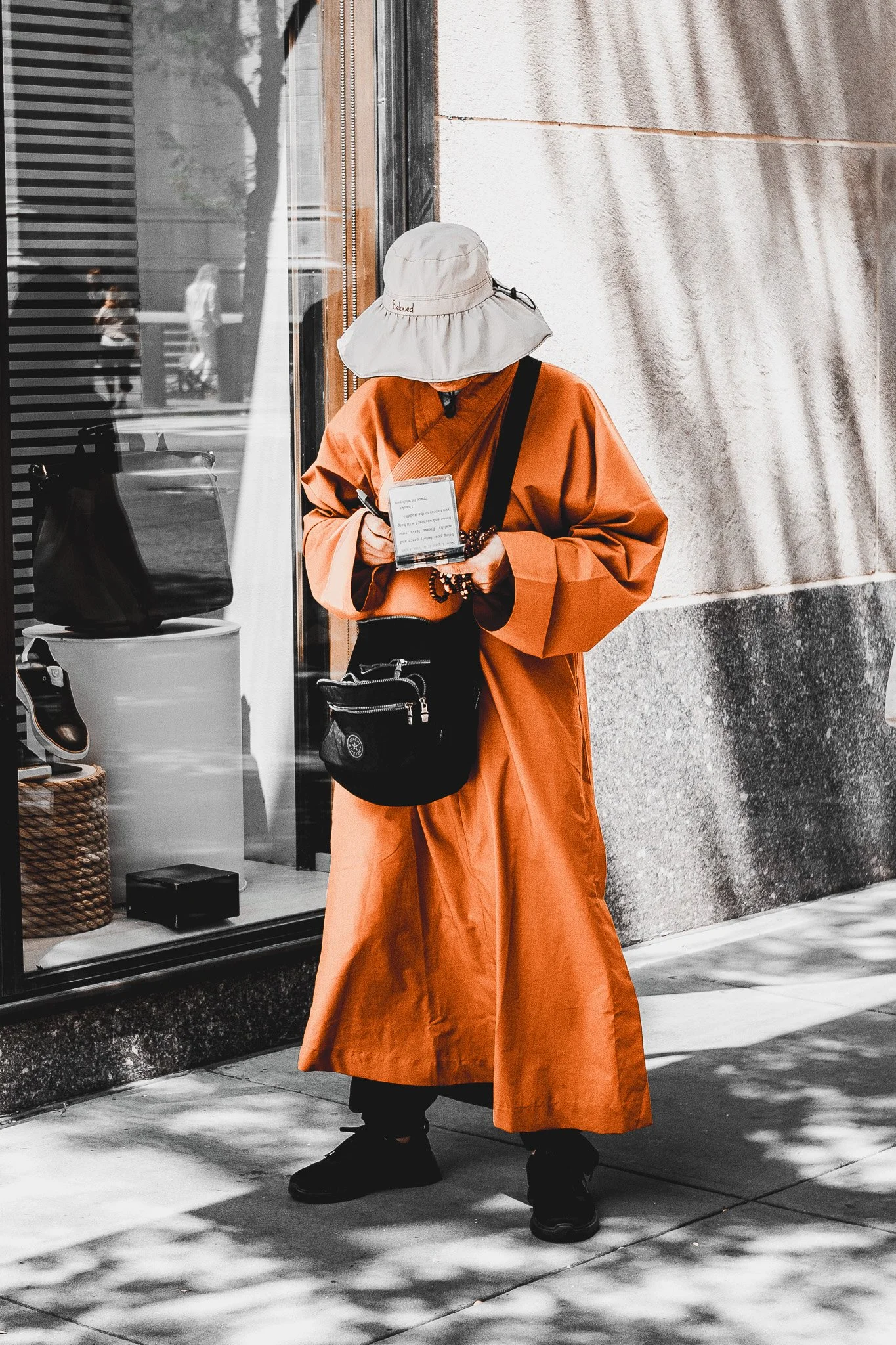 Person wearing a white bucket hat with word 'Beloved' embroidered on it, orange long coat, black boots, carrying a black crossbody bag, standing outside a storefront, holding a smartphone, with reflections of people and trees in the window.