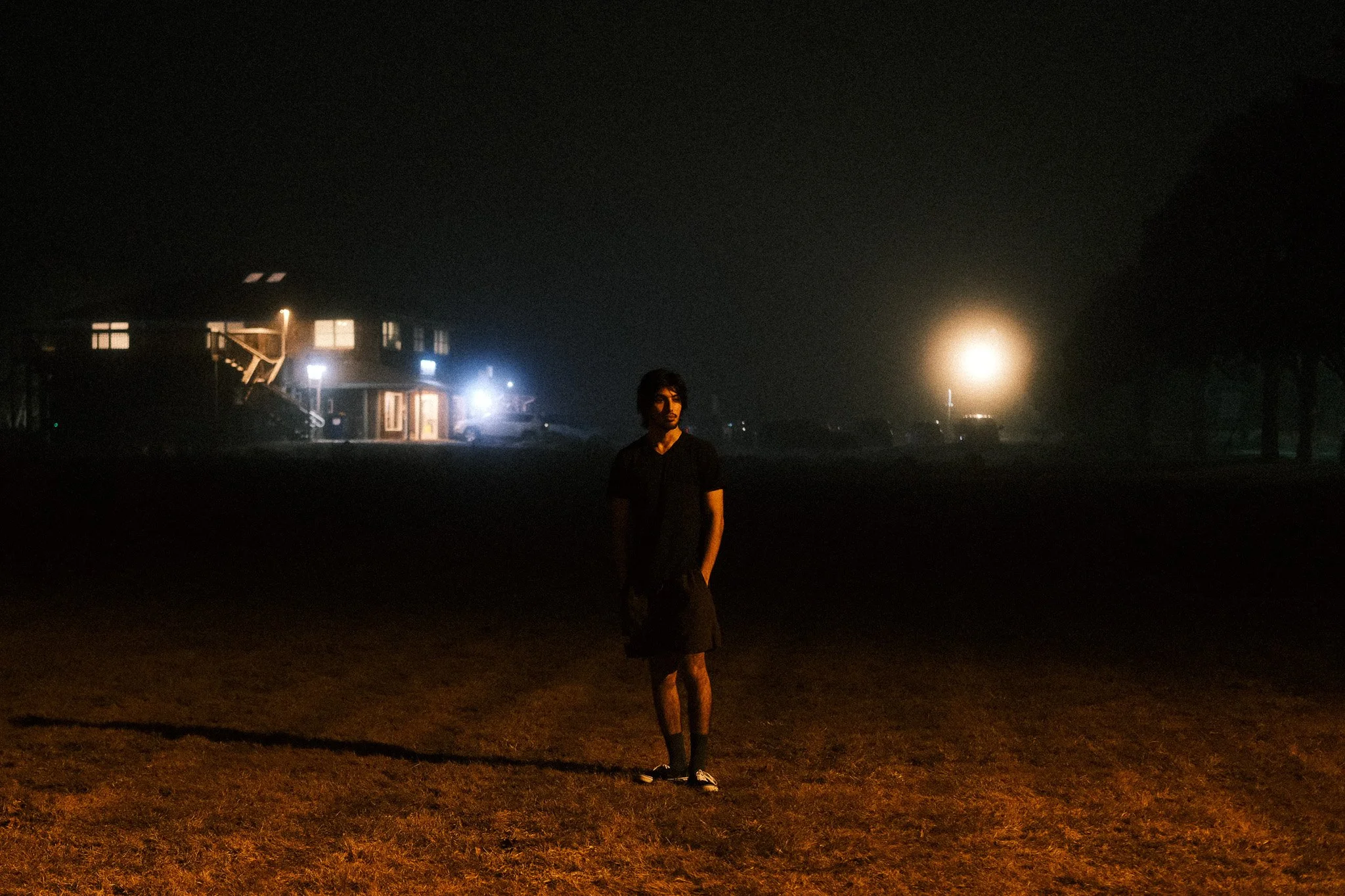 A person standing on a grassy area at night with houses, streetlights, and trees in the background, illuminated by bright lights.