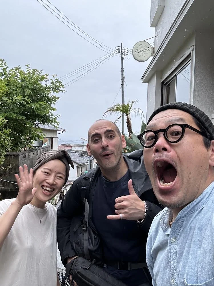 Three people taking a selfie outdoors in front of a house with a Sampar sign, with cloudy sky, power lines, a tree, and a palm tree in the background. They appear happy and excited.