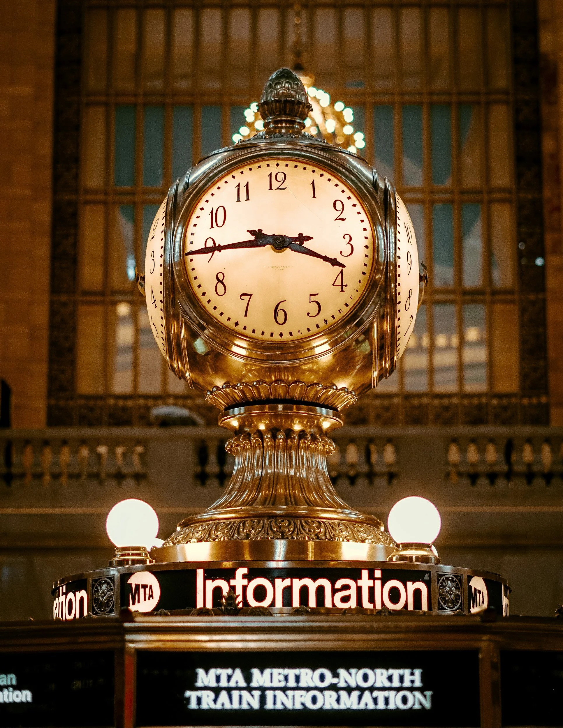 The Clock with an ornate gold frame and face showing the time as 3:45 at Grand Central Station, located above a digital train information display at a train station.