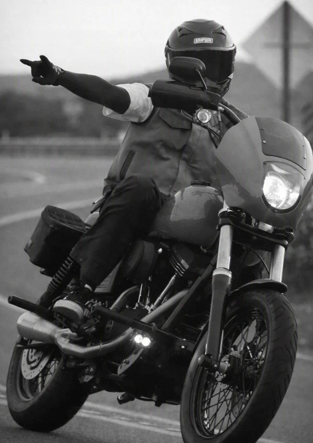 Motorcycle rider wearing a helmet and vest, with one arm extended outward, riding on an open road in black and white.