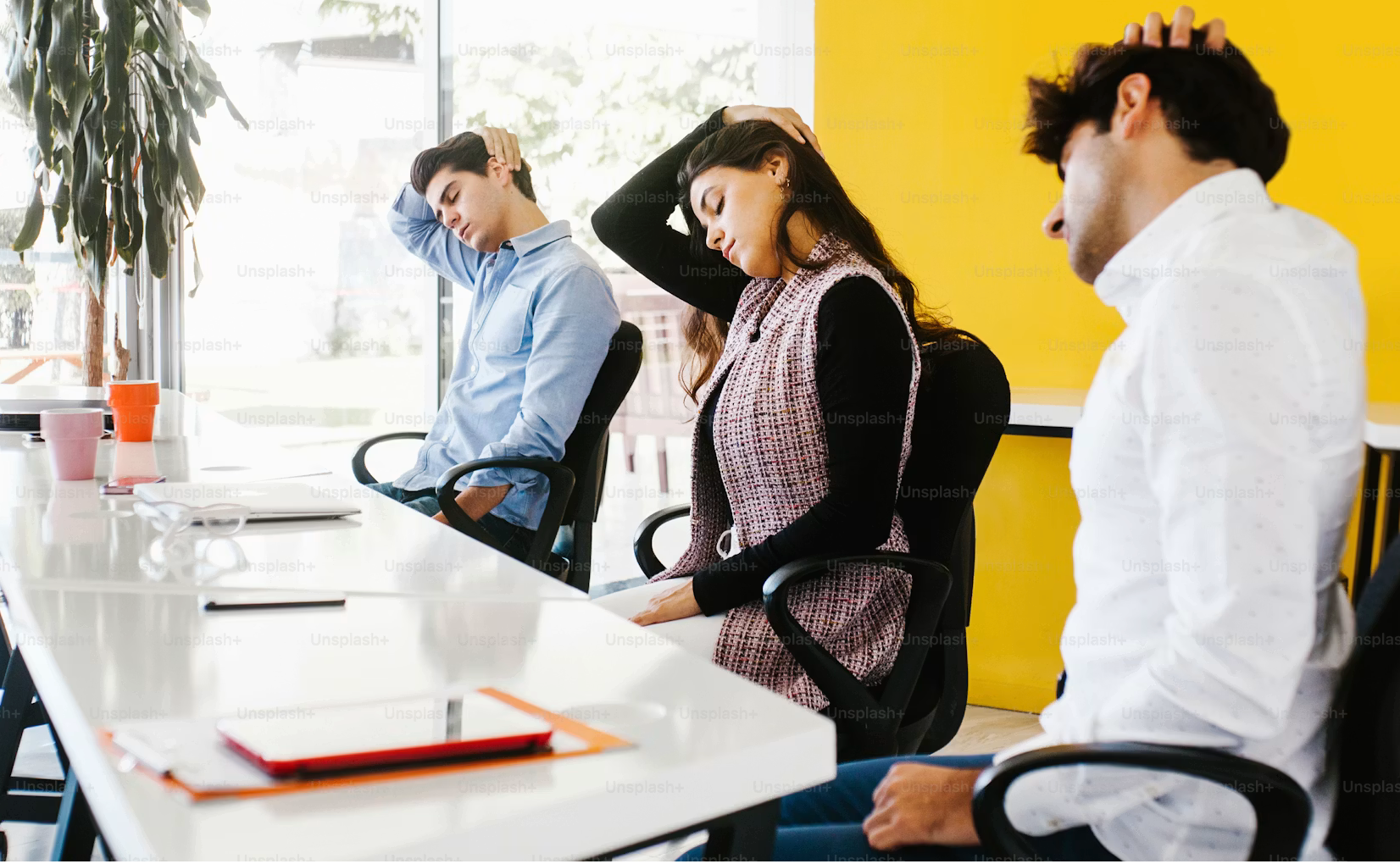 Three business people sitting at a conference table, visibly tired or stressed, with hands on their heads.