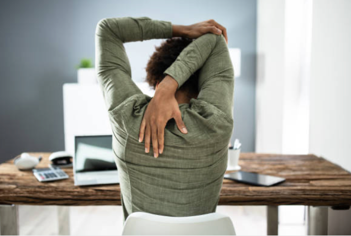 Person sitting at a desk stretching with arms behind head in an office setting.