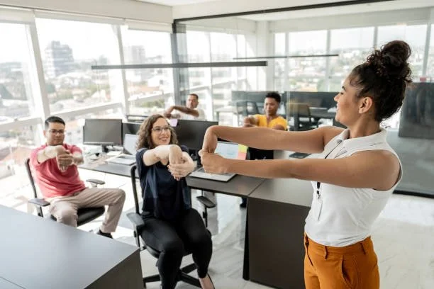 Group of coworkers in an office, sitting at desks with computers, making a fist bump with a woman standing and engaging with them.