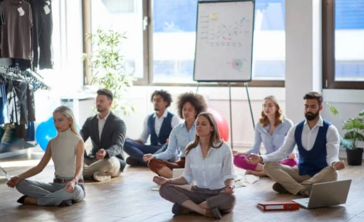 Group of people practicing meditation in a modern office with large windows, plants, and a whiteboard with notes.