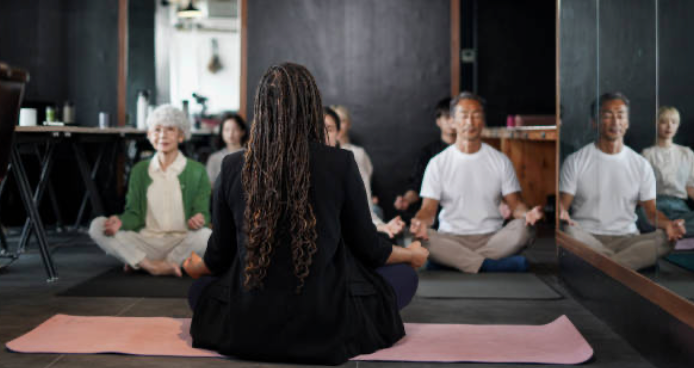 A diverse group of people practicing meditation in a yoga studio with a woman instructor at the front.