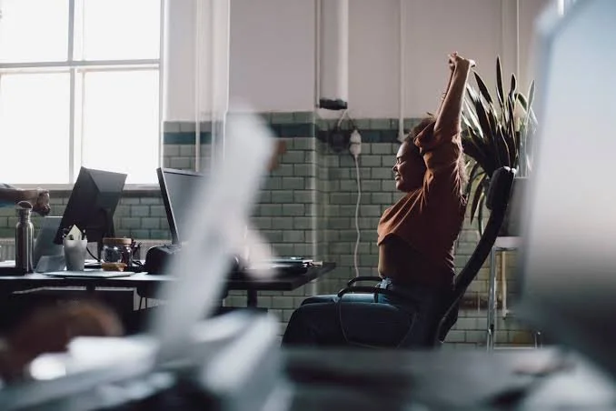 Woman sitting at her desk in an office stretching with arms raised after waking up or feeling tired.